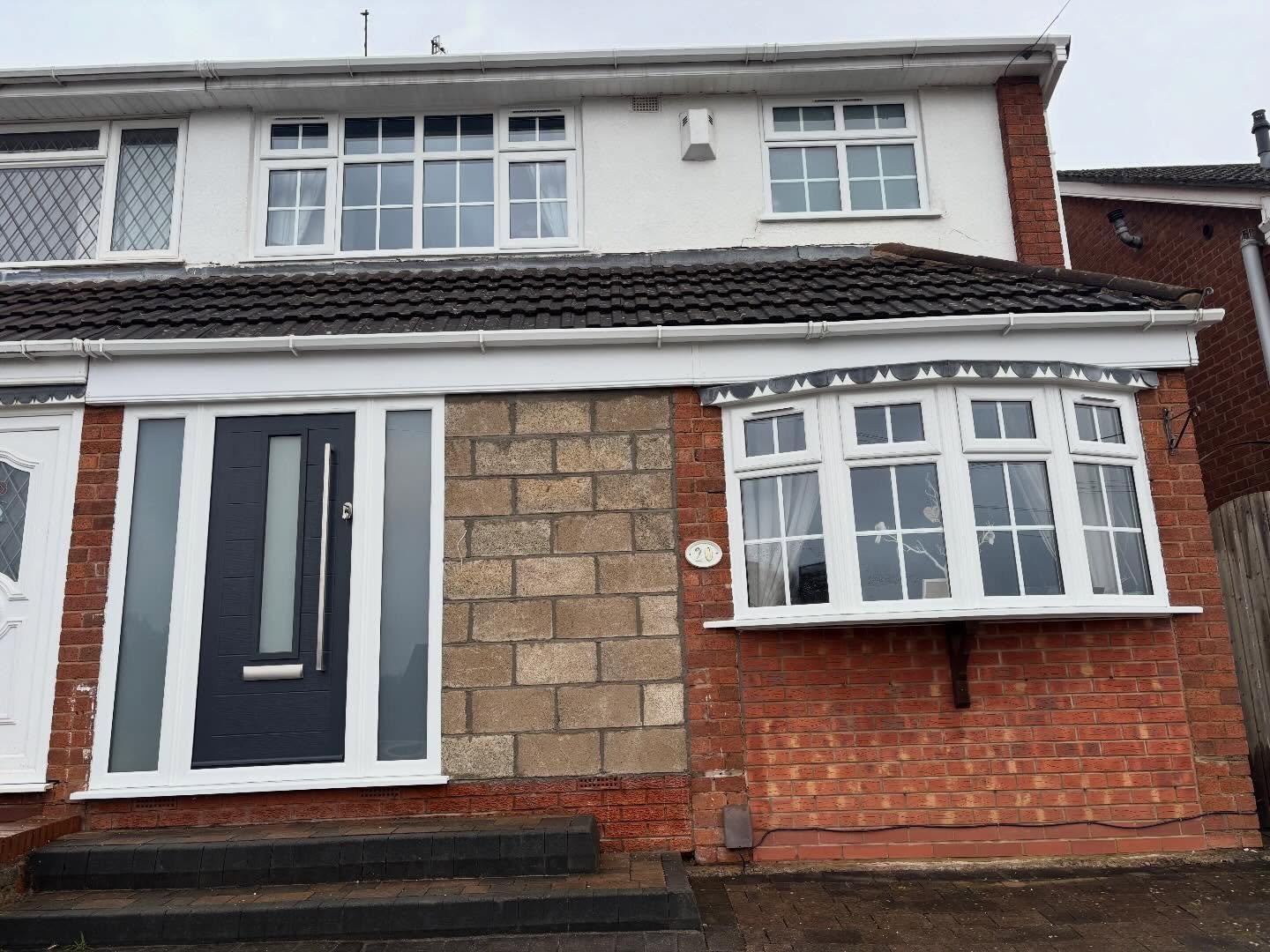Exterior of a two-story house with a front door, large bay window, and a mix of brick, stone, and white siding. The house has a black front door with vertical glass and a metallic handle, and the upper floor has multiple windows.