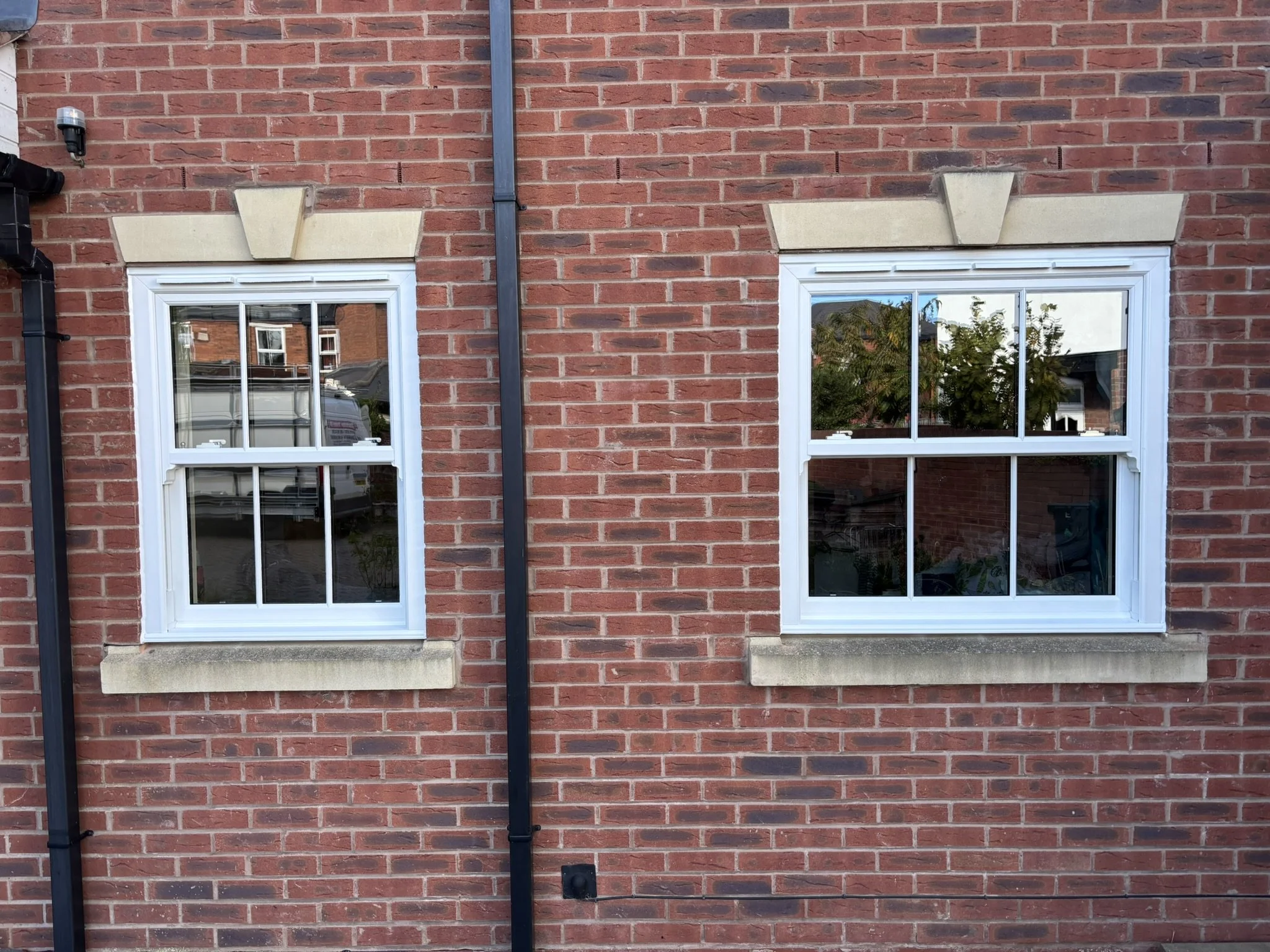 Two white-framed windows on a red brick wall, each with a stone lintel and sill, reflecting an outdoor scene including trees and nearby buildings, with a black drainpipe running vertically between them.