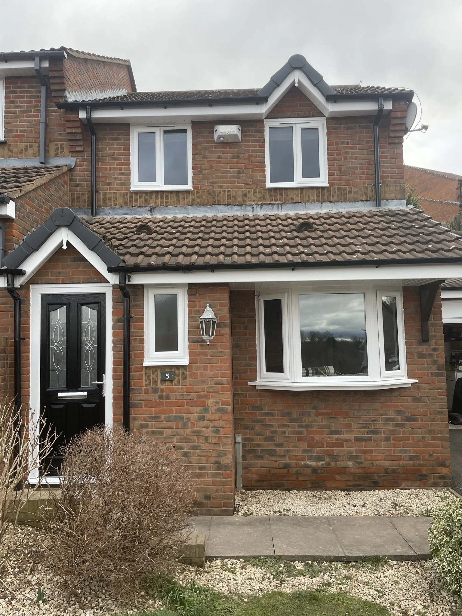 A two-story brick house with a black front door, white framed windows, a bay window, and a small front yard with gravel and bushes.