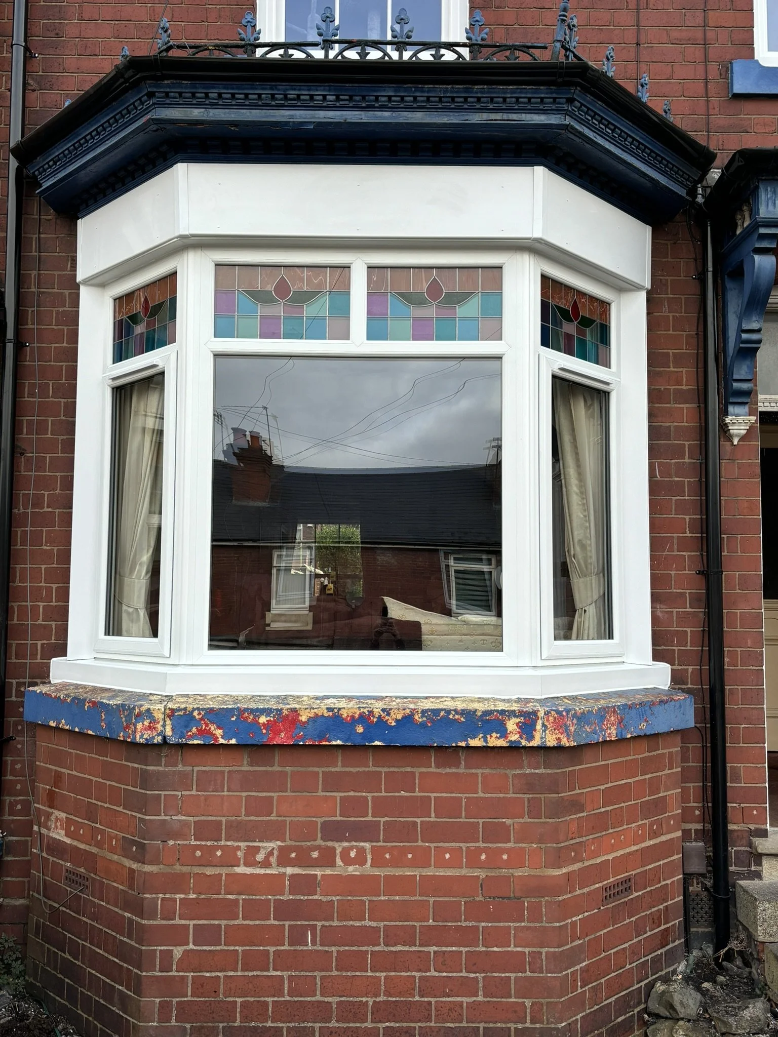 A brick house with a large bay window decorated with stained glass at the top and cream curtains inside. The window has white framing and a level blue ledge with faded, chipped paint. The house features dark blue trim and decorative metalwork on top 