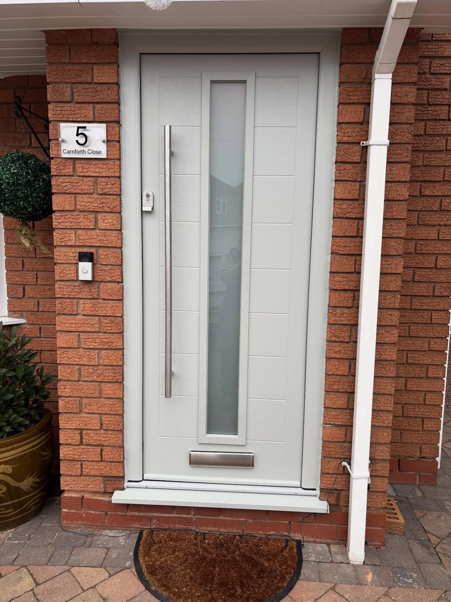 White front door with a vertical frosted glass window, set in a red brick house wall, with house number 5 and a street name sign 'Carnforth Close' to the side.
