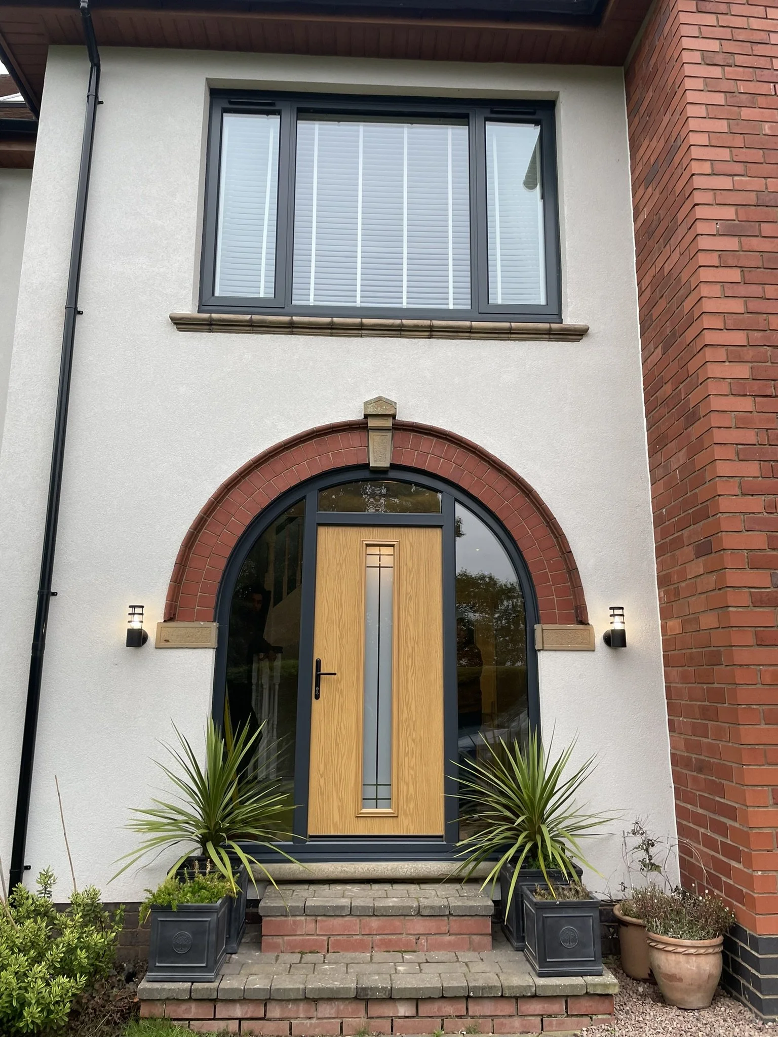 Front entrance of a house with an arched brick frame, a wooden front door, stairs, and potted plants.
