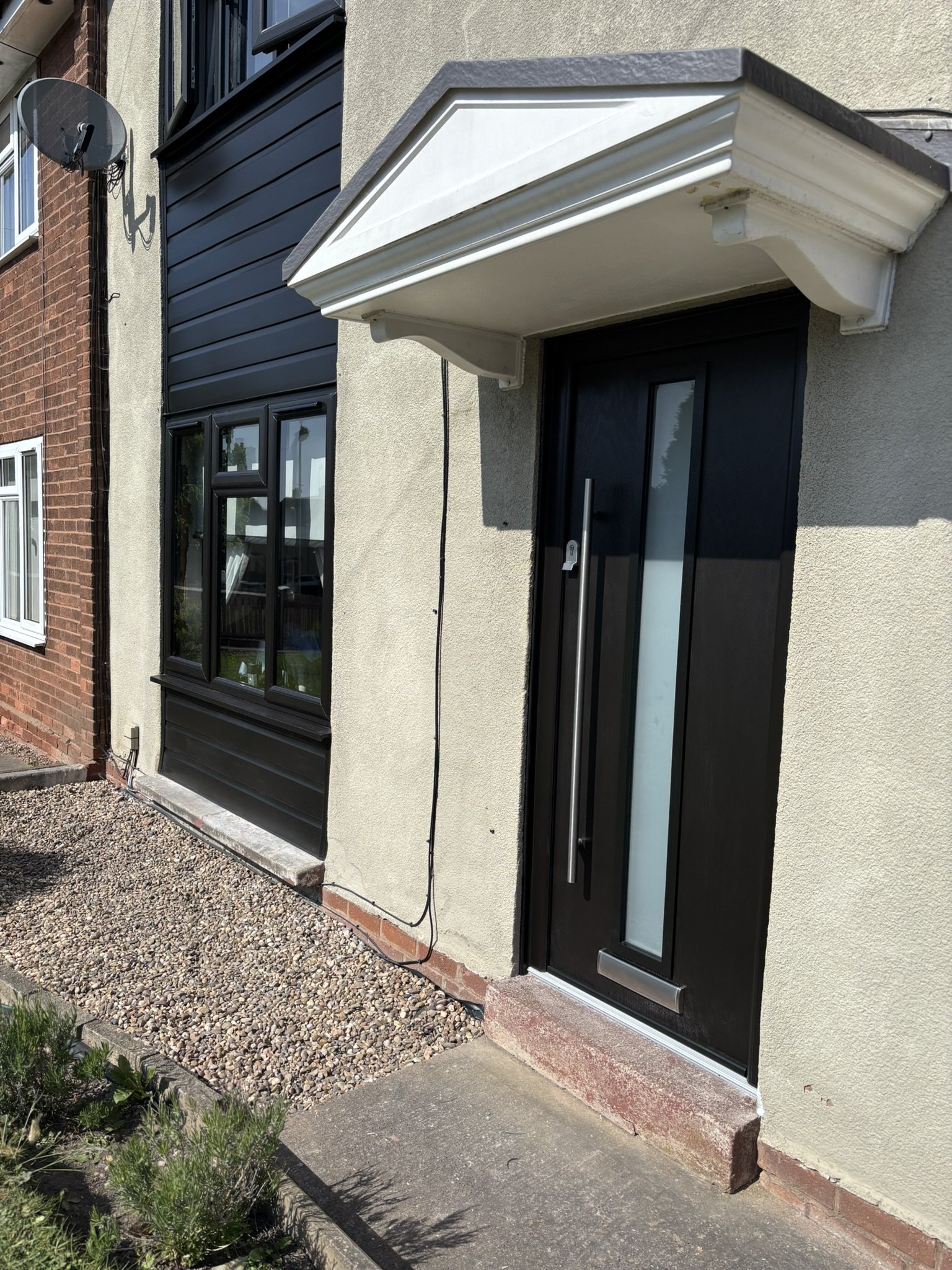 Close-up of a black front door with a vertical window pane, silver handle, mailbox, and a white decorative porch overhang. The house has beige walls and neighboring brick and black-paneled exteriors.