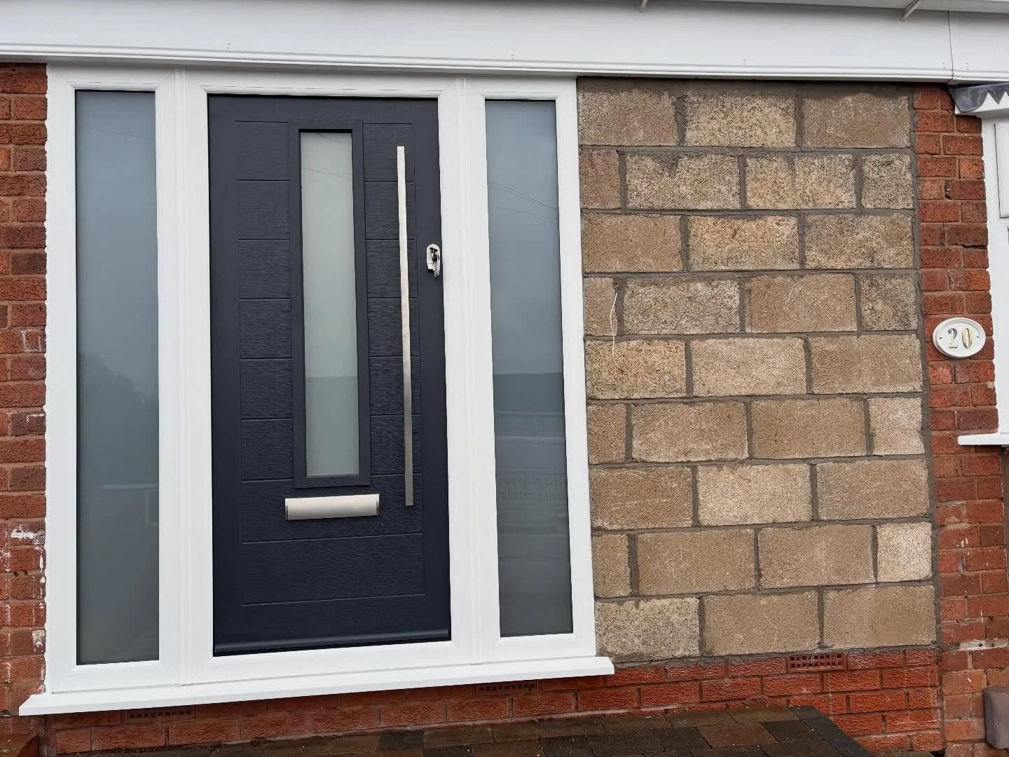 Front door to a house with a black door, frosted glass panels, and a silver handle, flanked by white-framed side and top windows, with a brick exterior and house number 20 on a white oval plaque.