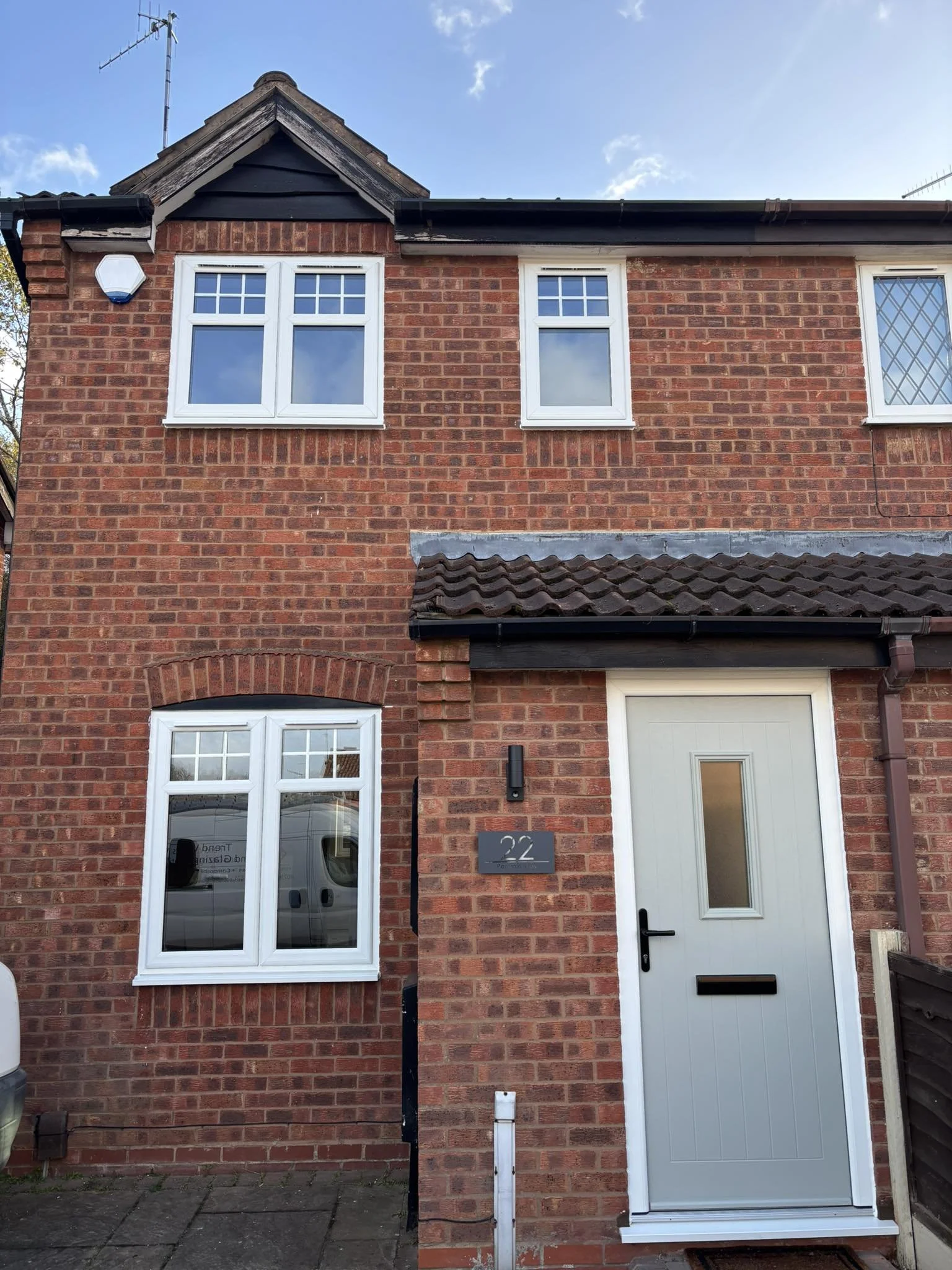 A brick two-story house with three white-framed windows on the second floor, one with a diamond-patterned window, & a window on the ground floor next to a gray front door. A house number 22 and a doorbell are visible beside the door. There's a drivew