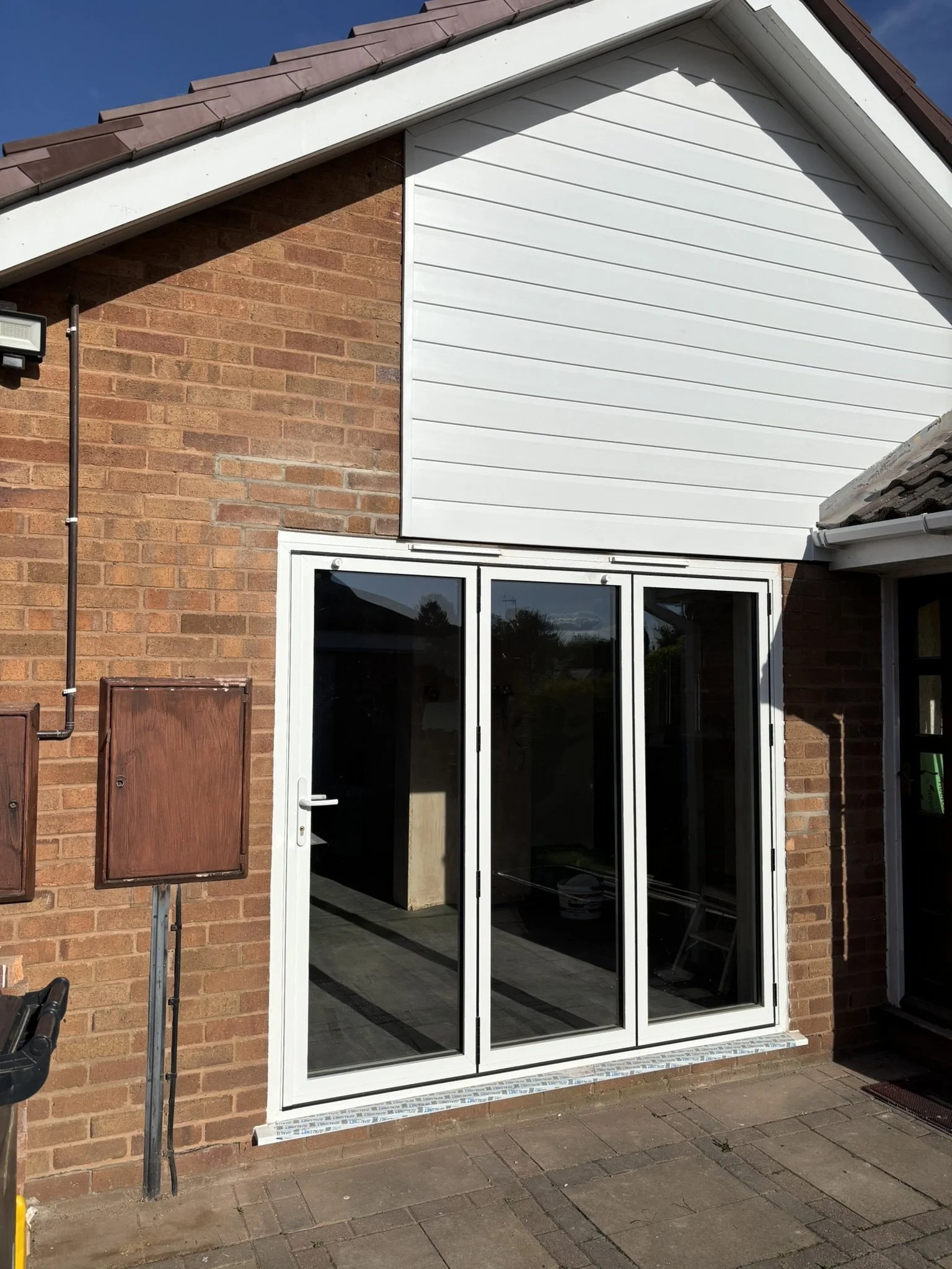 Exterior view of a house with brick walls and a sloped roof, featuring a large glass sliding door, a white upper section siding, and some utility boxes and pipes outside.