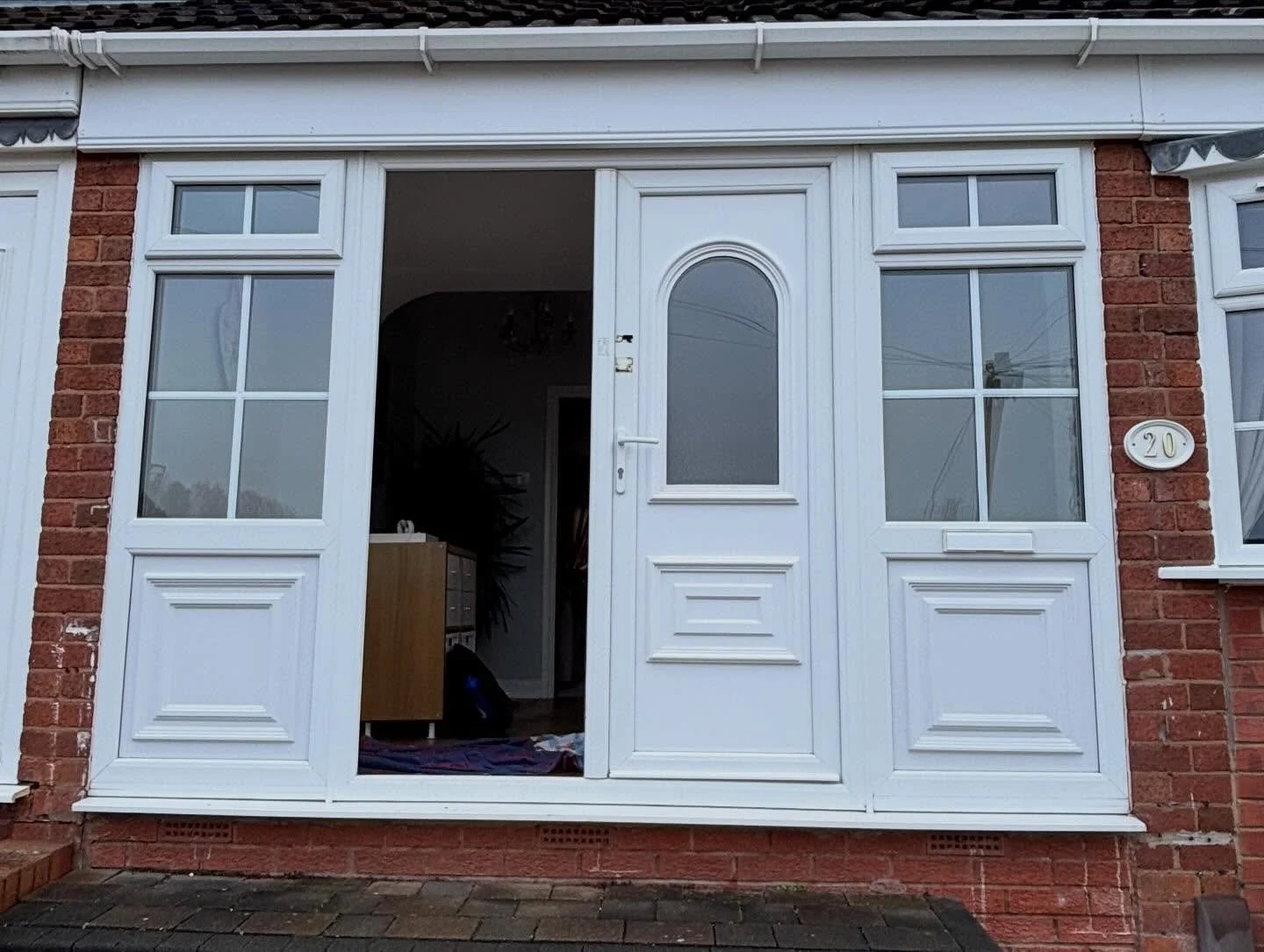 Front view of a house with a white porch enclosure, featuring a door with an arched window and house number 20, brick walls on either side, and large windows with divided panes.
