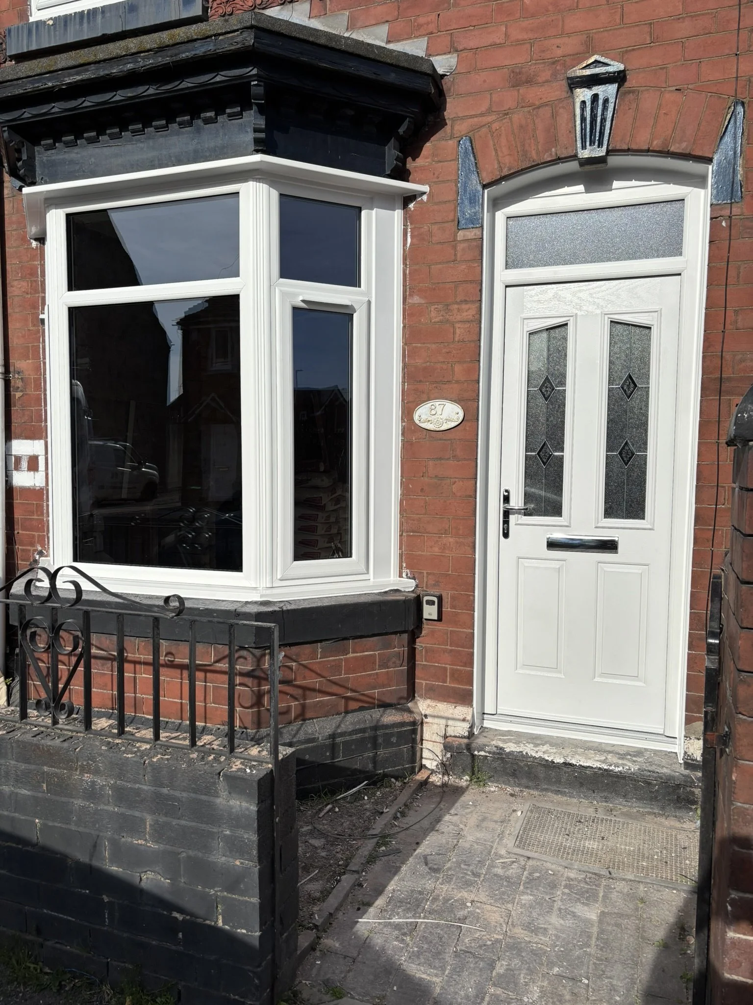 Brick house with a white door and a large white-framed bay window, small front garden area with a black metal fence, house number 87 visible next to door, and decorative wall fixtures above door and window.