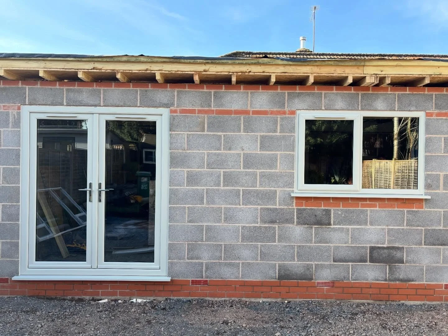 A partially constructed building with gray brick walls, red brick accents, a glass double door, and a window, under a roof with exposed wooden beams and black roofing material. There is an antenna on the roof, and the ground is gravel.