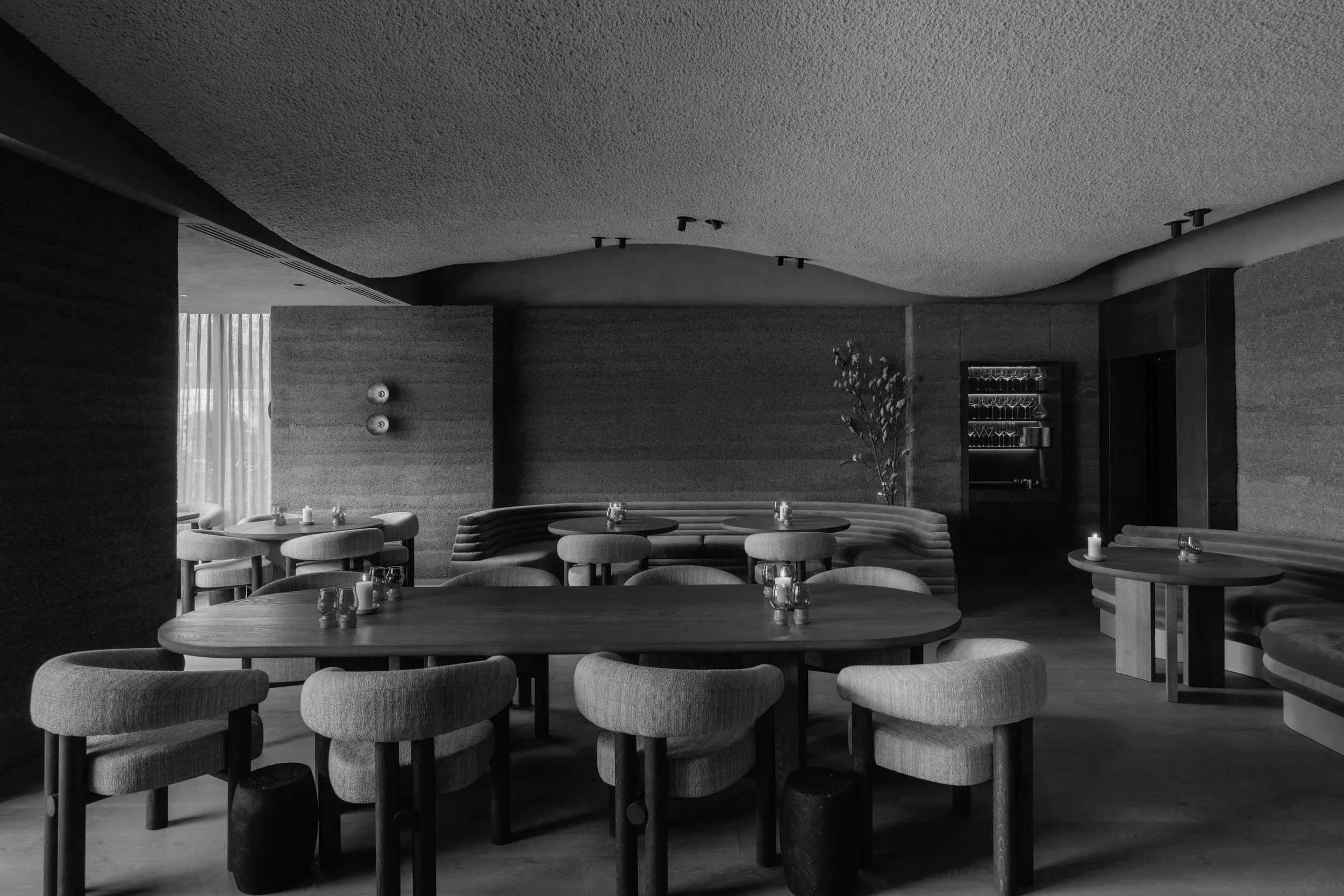 A modern indoor dining area with round tables, upholstered chairs, and soft lighting, featuring a minimalist design with dark textured walls and ceiling.