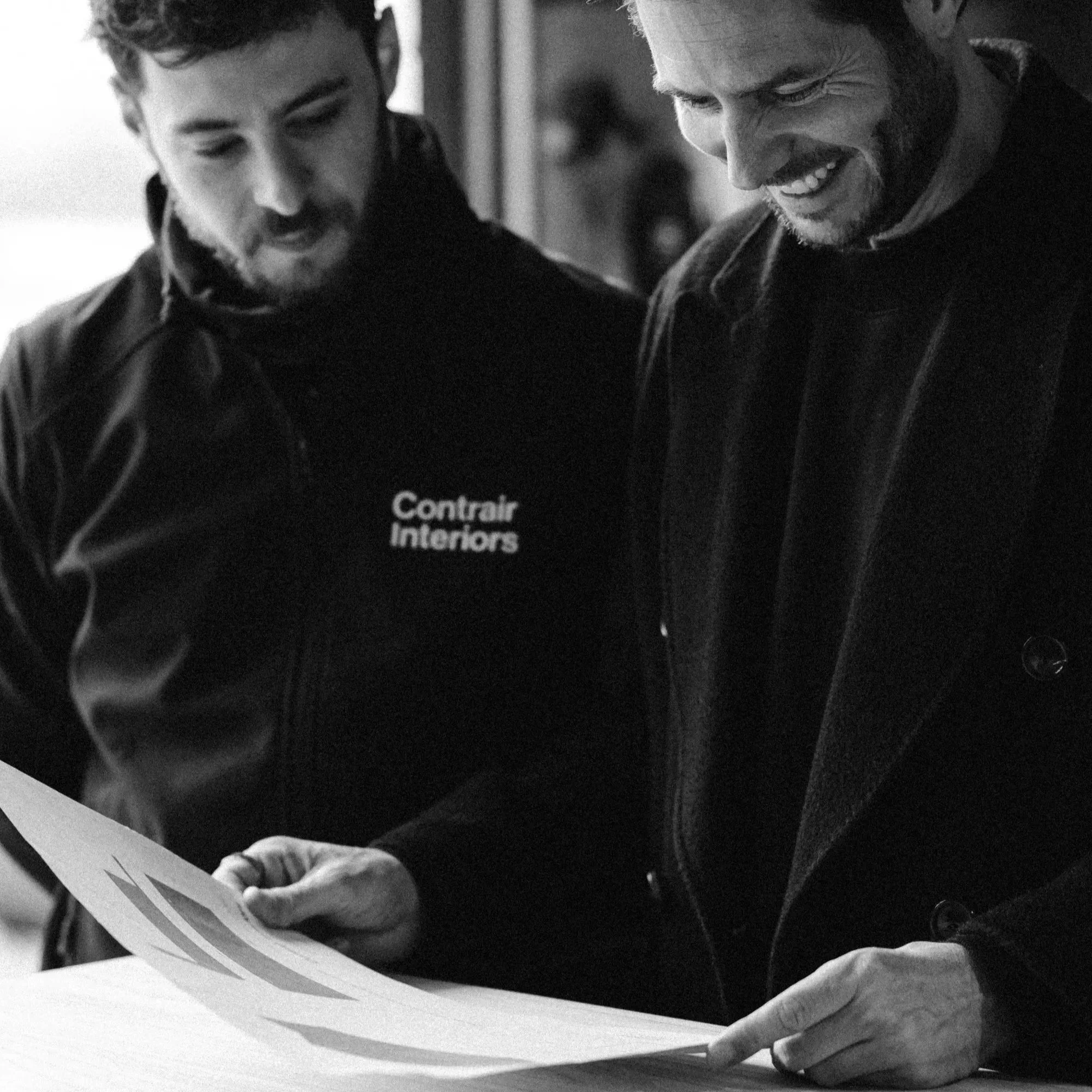 Two men are inspecting a constructing plan. One man is wearing a jacket with the words 'Contrair Interiors' on it, and the other man is smiling and looking down at the model.