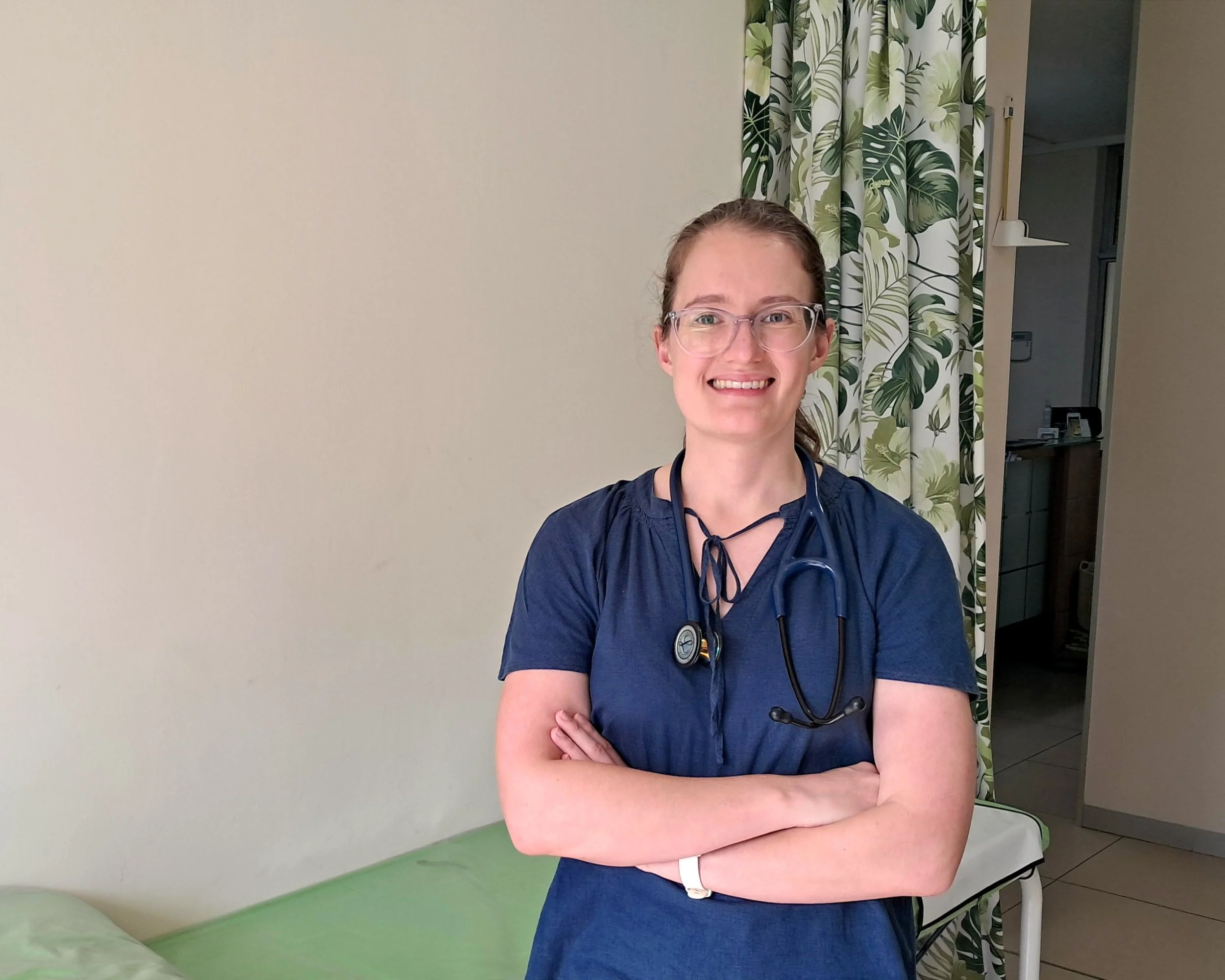 A smiling female healthcare professional with glasses and a stethoscope around her neck, standing in a hospital room.