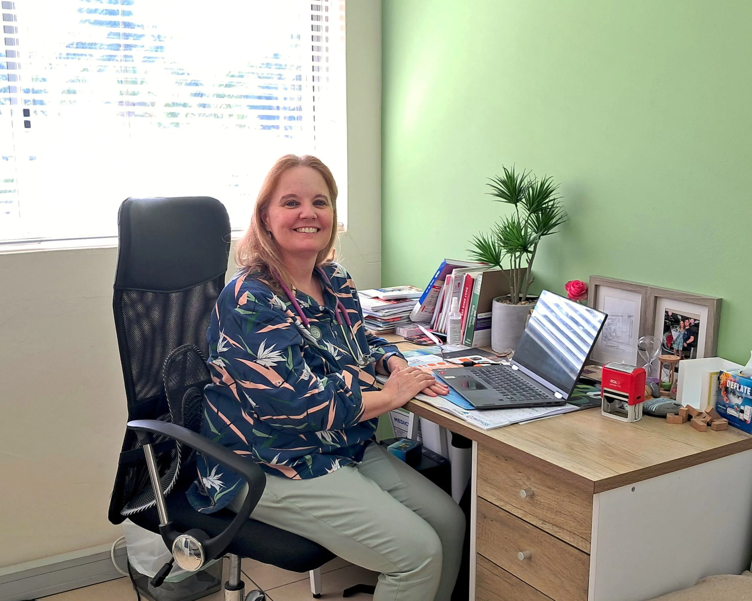 A woman in medical scrubs sitting at a cluttered desk in an office with green and white walls, smiling at the camera.