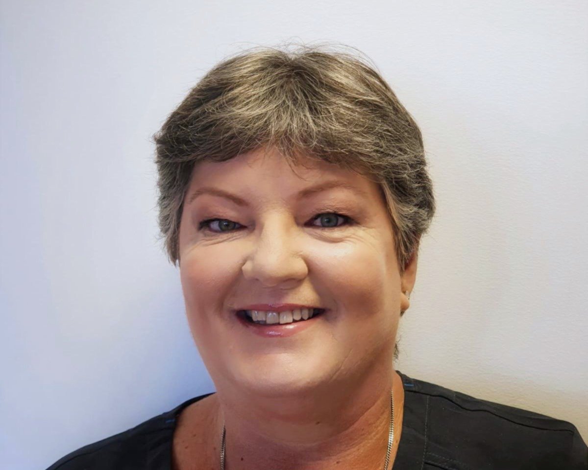 Close-up portrait of a smiling woman with short gray hair, light skin, and wearing a black top and a necklace, standing against a plain light-colored background.