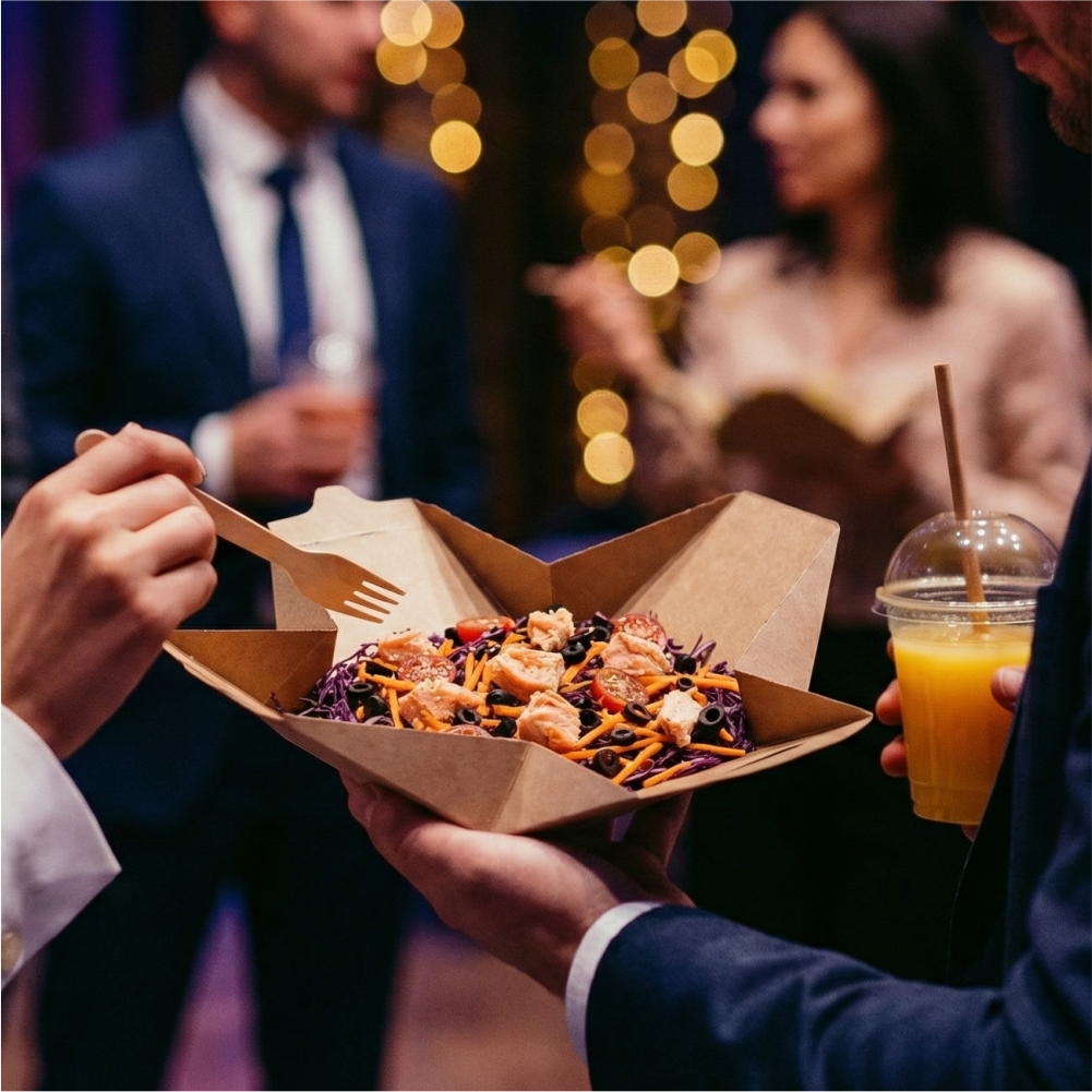 People at a social gathering holding a tray with a colorful salad, while one person is holding a drink, with blurred background lights and two other people in conversation.