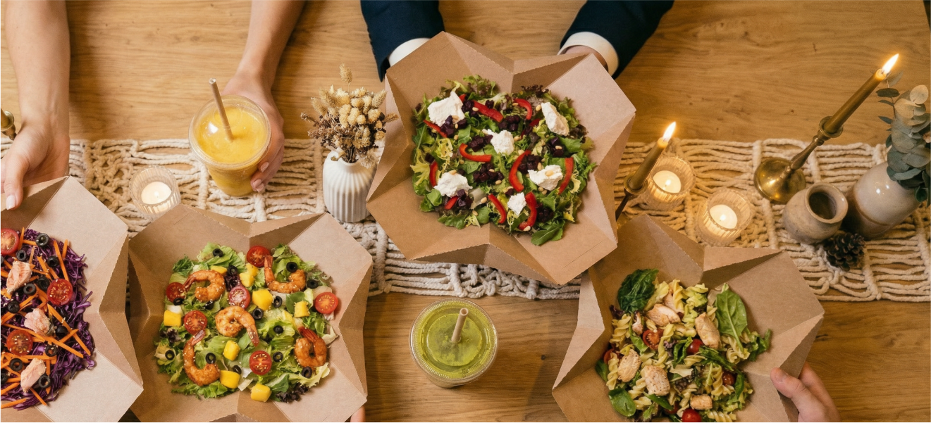 Various salads in takeout containers on a wooden table with candles, a small vase of dried flowers, and four people reaching for food.