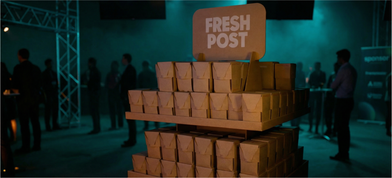 Stack of brown paper food boxes under a sign that says 'FRESH POST' at an event with people in the background.