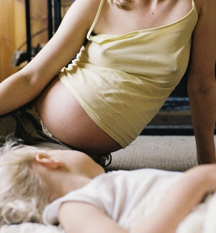 A pregnant woman sitting on the floor with a young child lying next to her, in a cozy indoor setting.