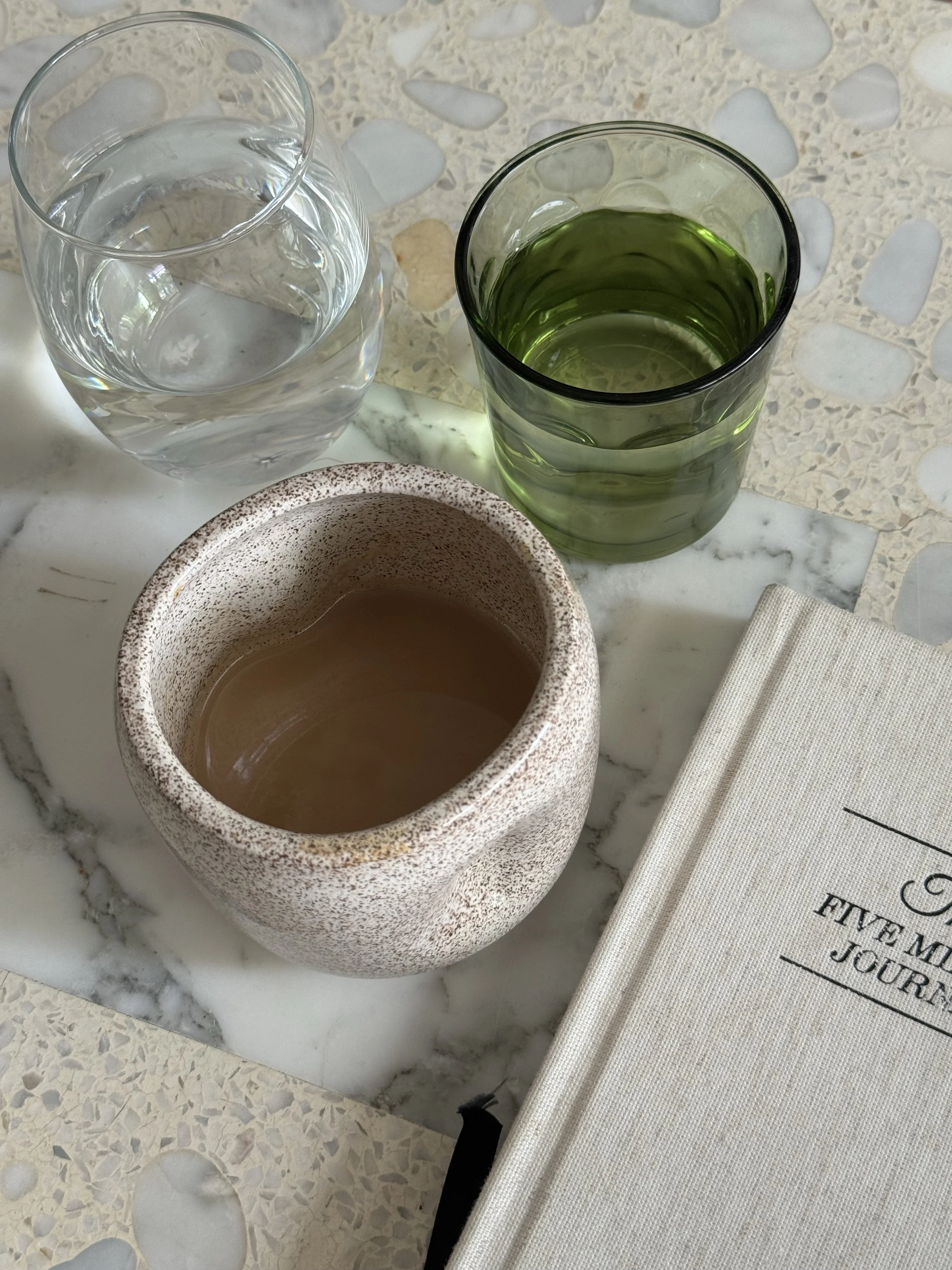 A marble table holding three drinks: a glass of water, a glass of green juice, and a ceramic mug with hot coffee, next to a book titled 'Five Minute Journal'.