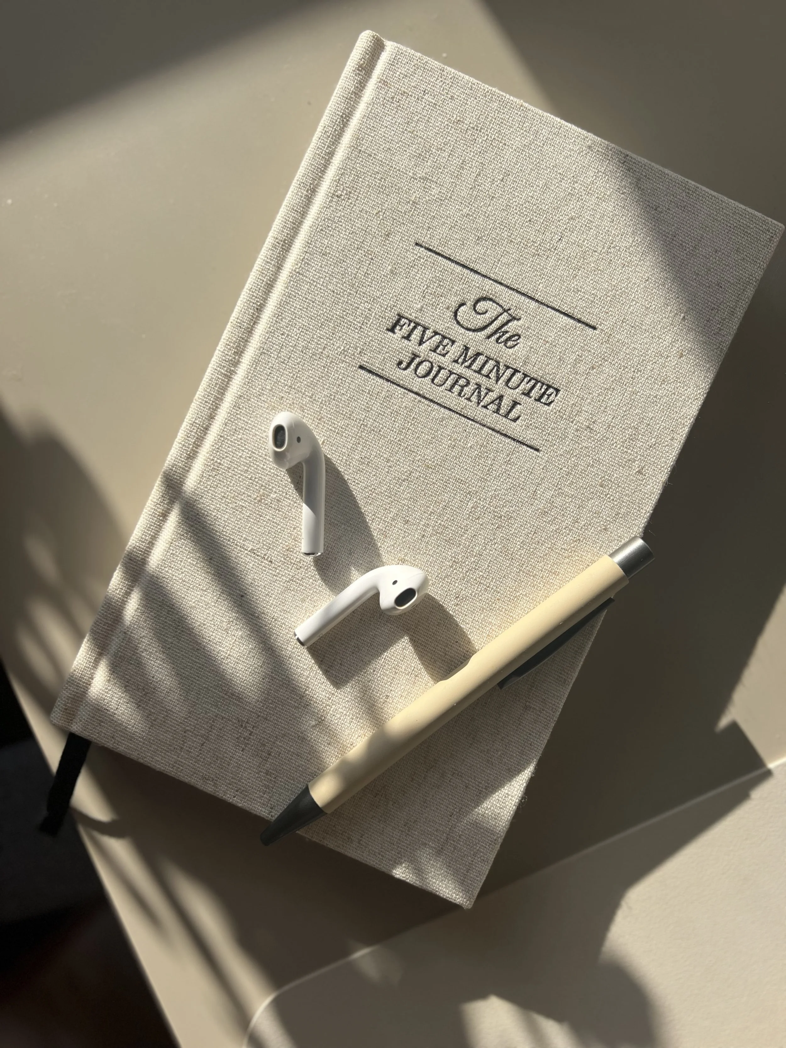 A beige fabric journal titled "The Five Minute Journal" with a black ribbon bookmark, a pair of white wireless earbuds, and a beige or light-colored pen with a silver tip resting on top. The journal is placed on a white surface, with sunlight casting shadows.