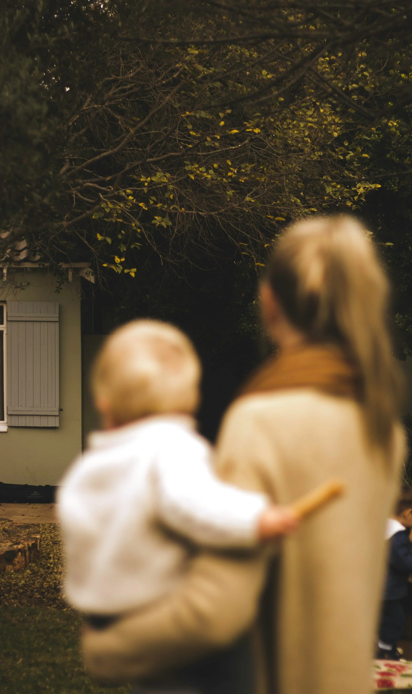 A woman holding a young blonde child with a blurred house and tree with yellow leaves in the background.