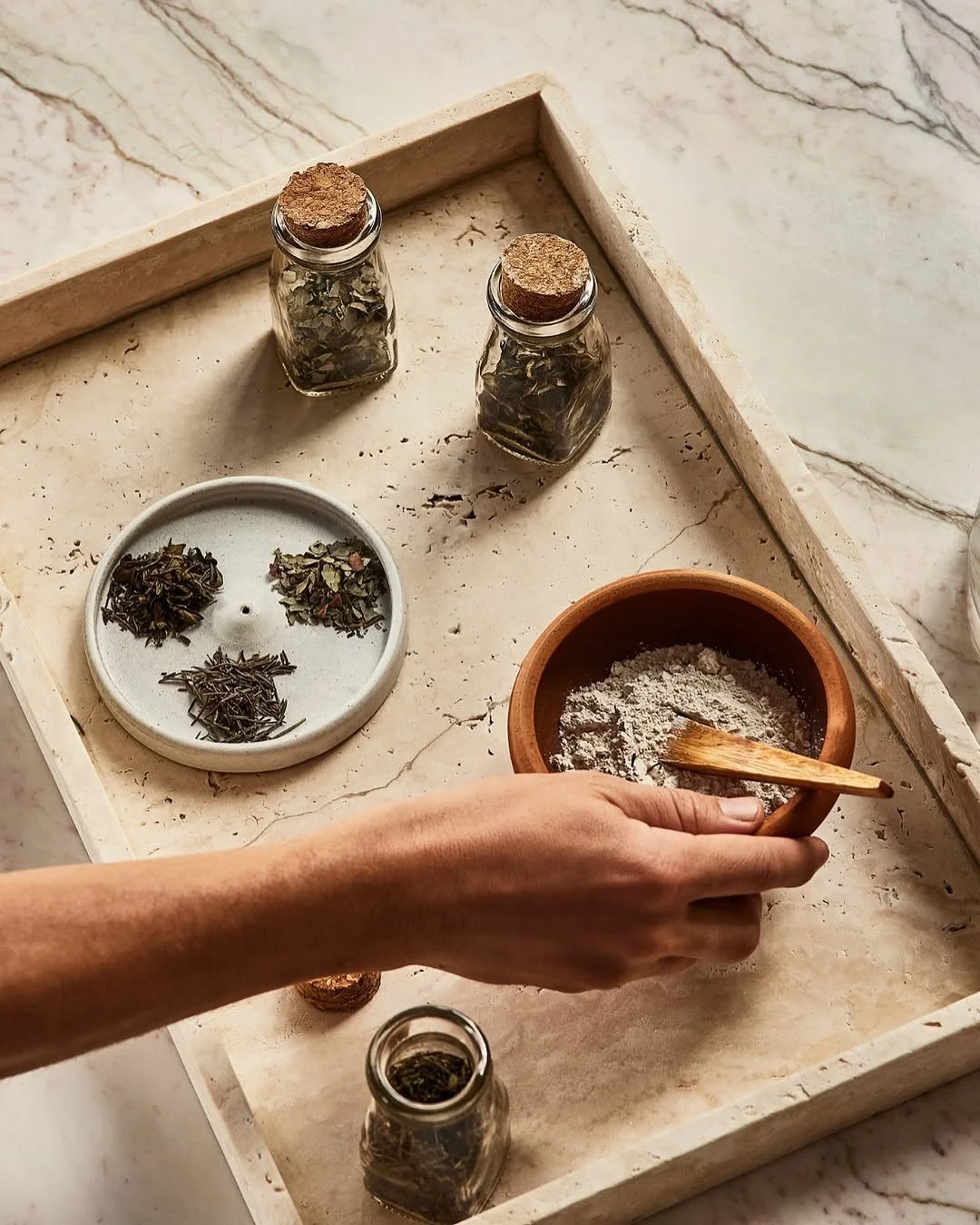 A person stirring powder in a clay bowl with a wooden spoon on a marble surface. Surrounding items include glass jars with dried herbs and a white dish with assorted herbs.