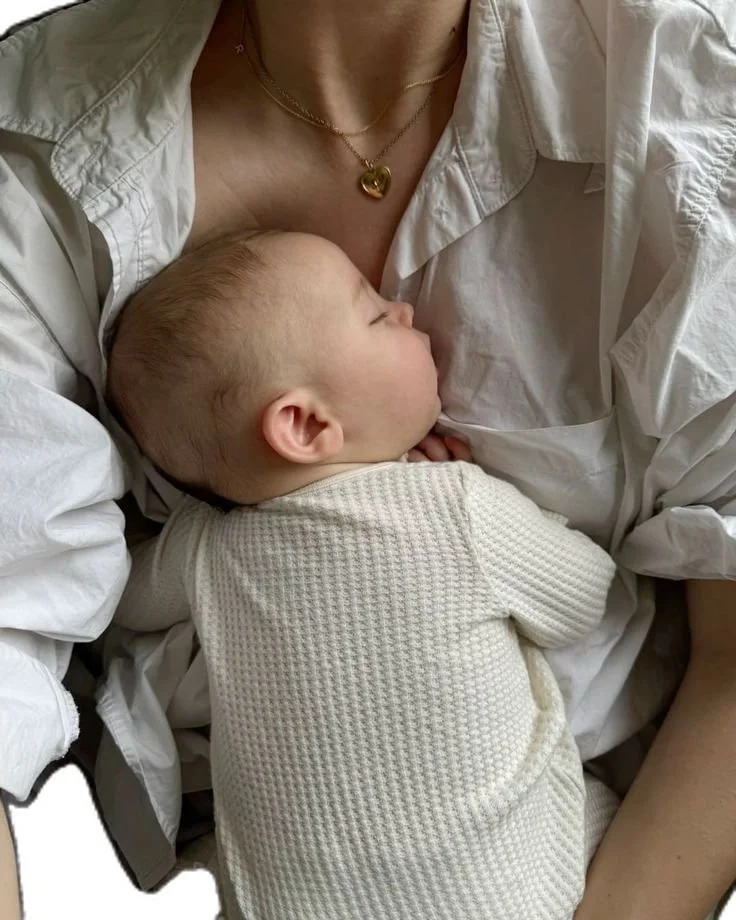 A person wearing a white shirt with a gold heart necklace holding a sleeping baby bundled in a cream knit blanket.