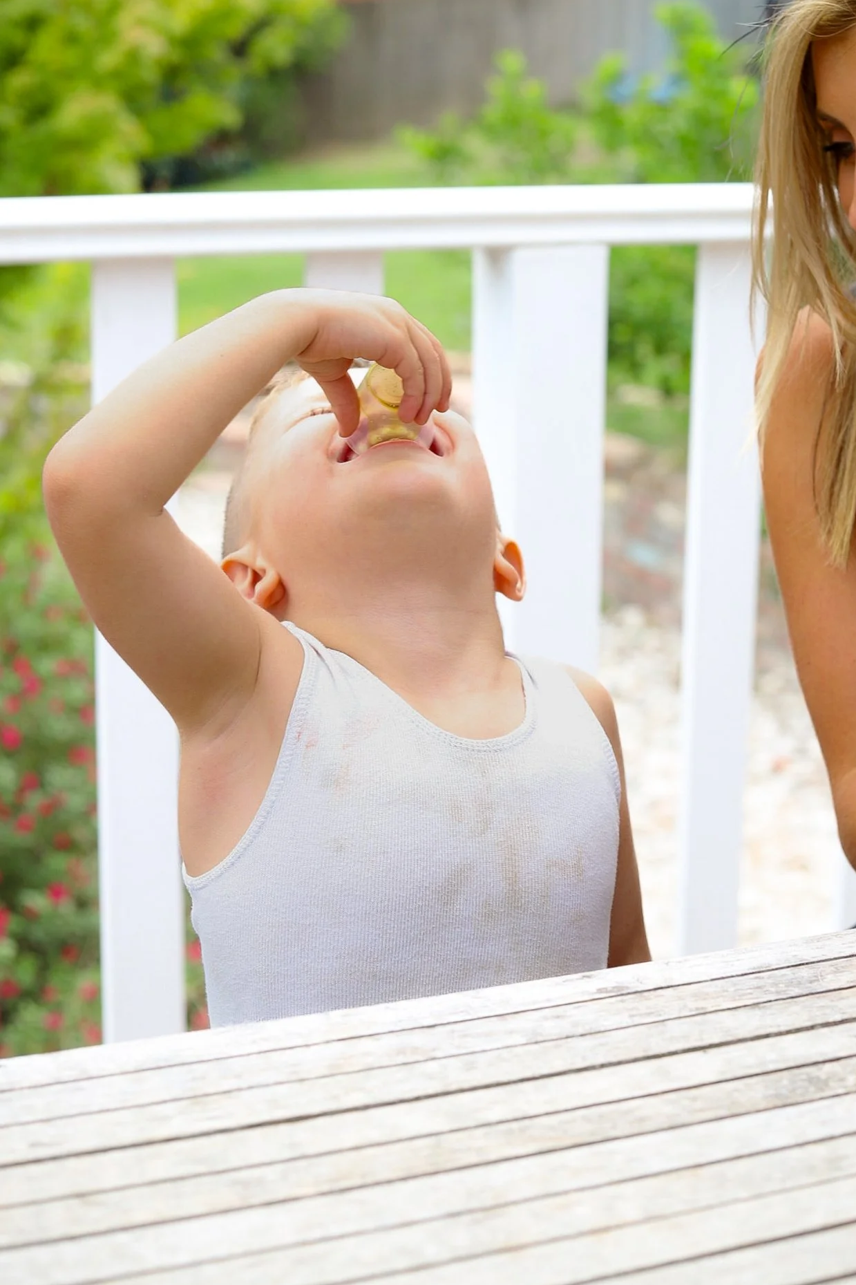 A young boy in a white tank top is outdoors, tilting his head back and drinking from a cup, with a woman partially visible next to him.