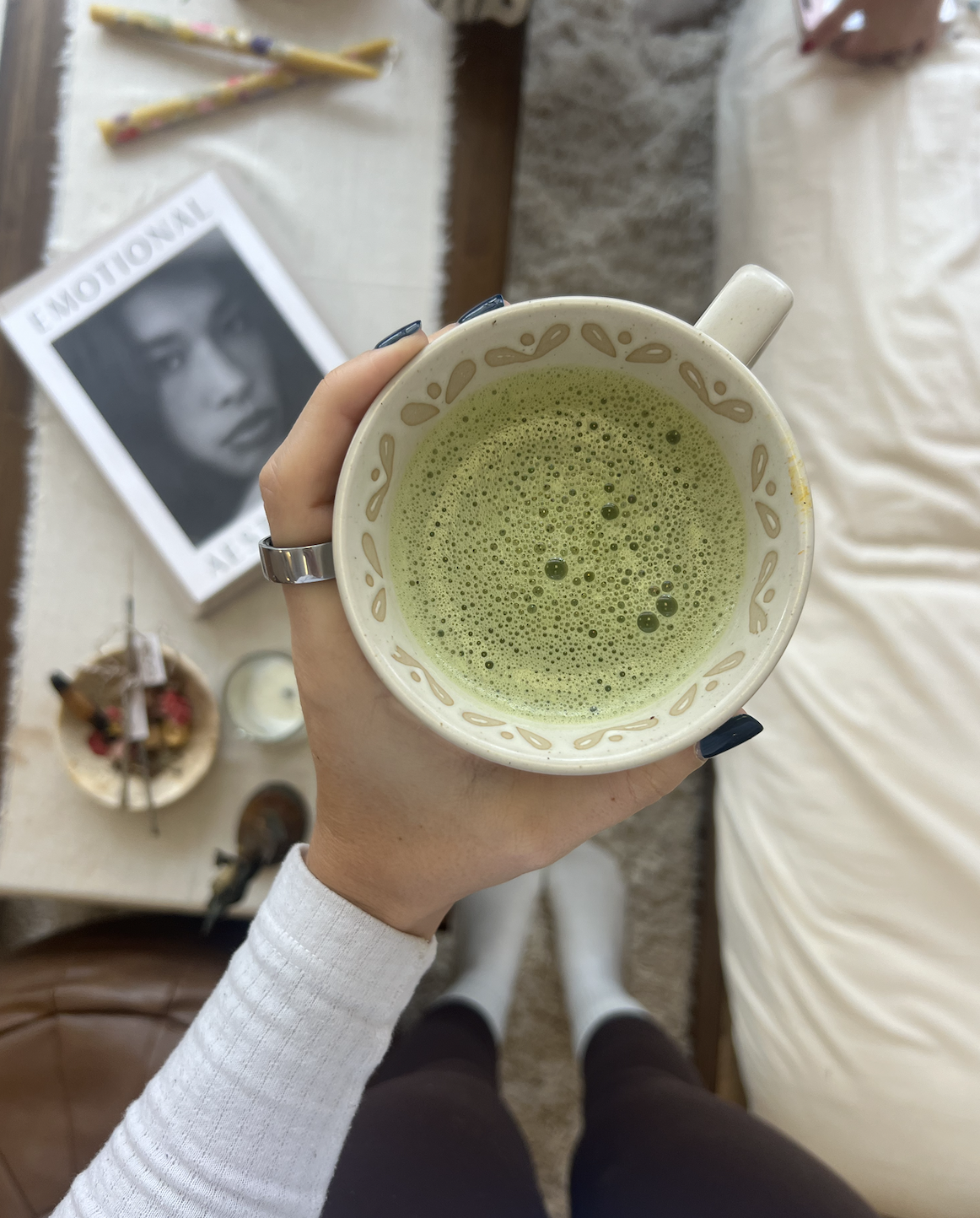 A person holding a white cup of frothy matcha tea with a decorative pattern on the rim, taking a selfie from above in a cozy room. A magazine, some snack trays, and other items can be seen on a table in the background.