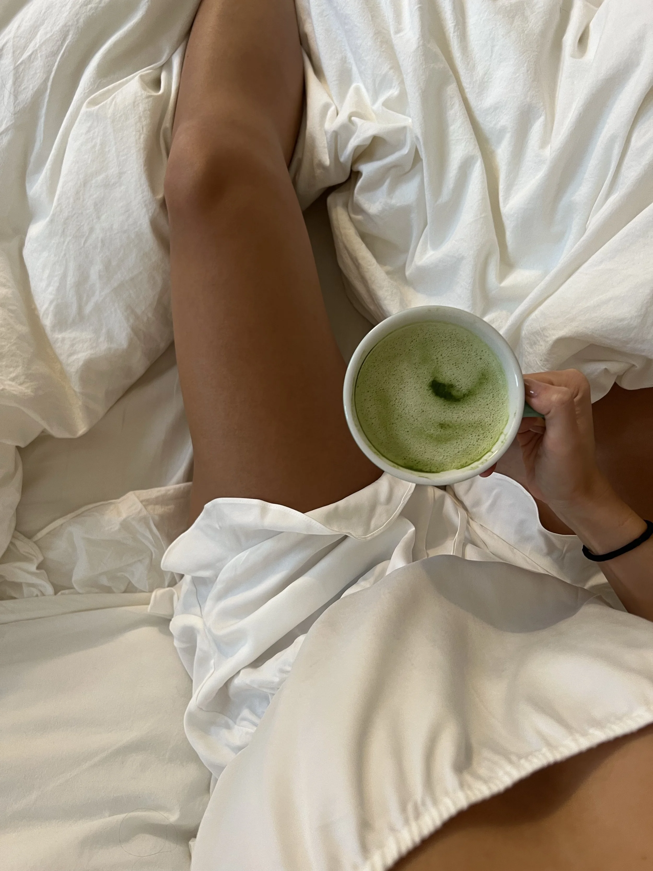Person sitting on a bed, holding a white mug with green tea or matcha, surrounded by white bed linens.