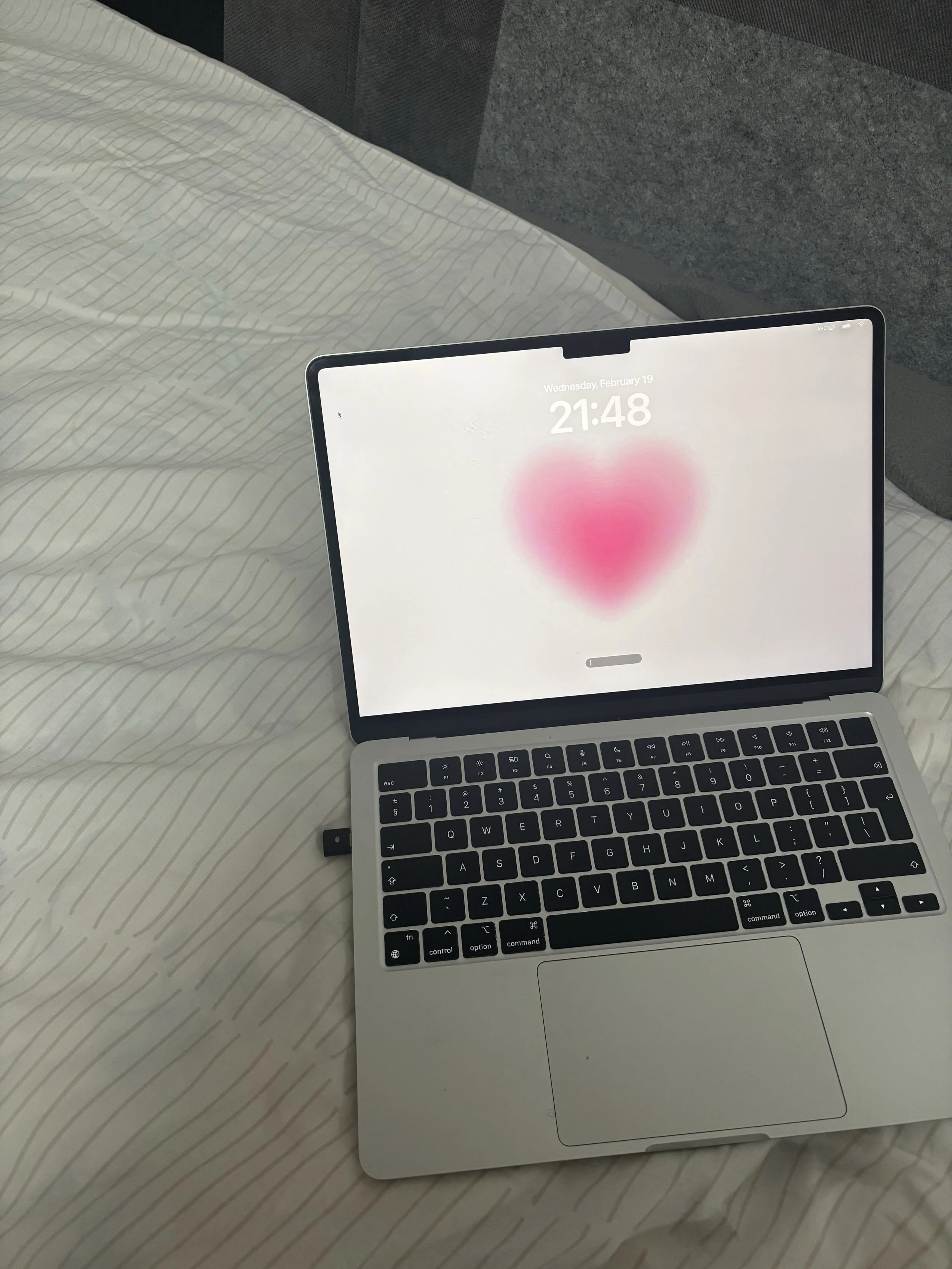 A laptop with a white body and black keyboard resting on a bed with white and beige striped sheets. The screen displays a blurred pink heart and the date and time, Wednesday, February 19 at 21:48.