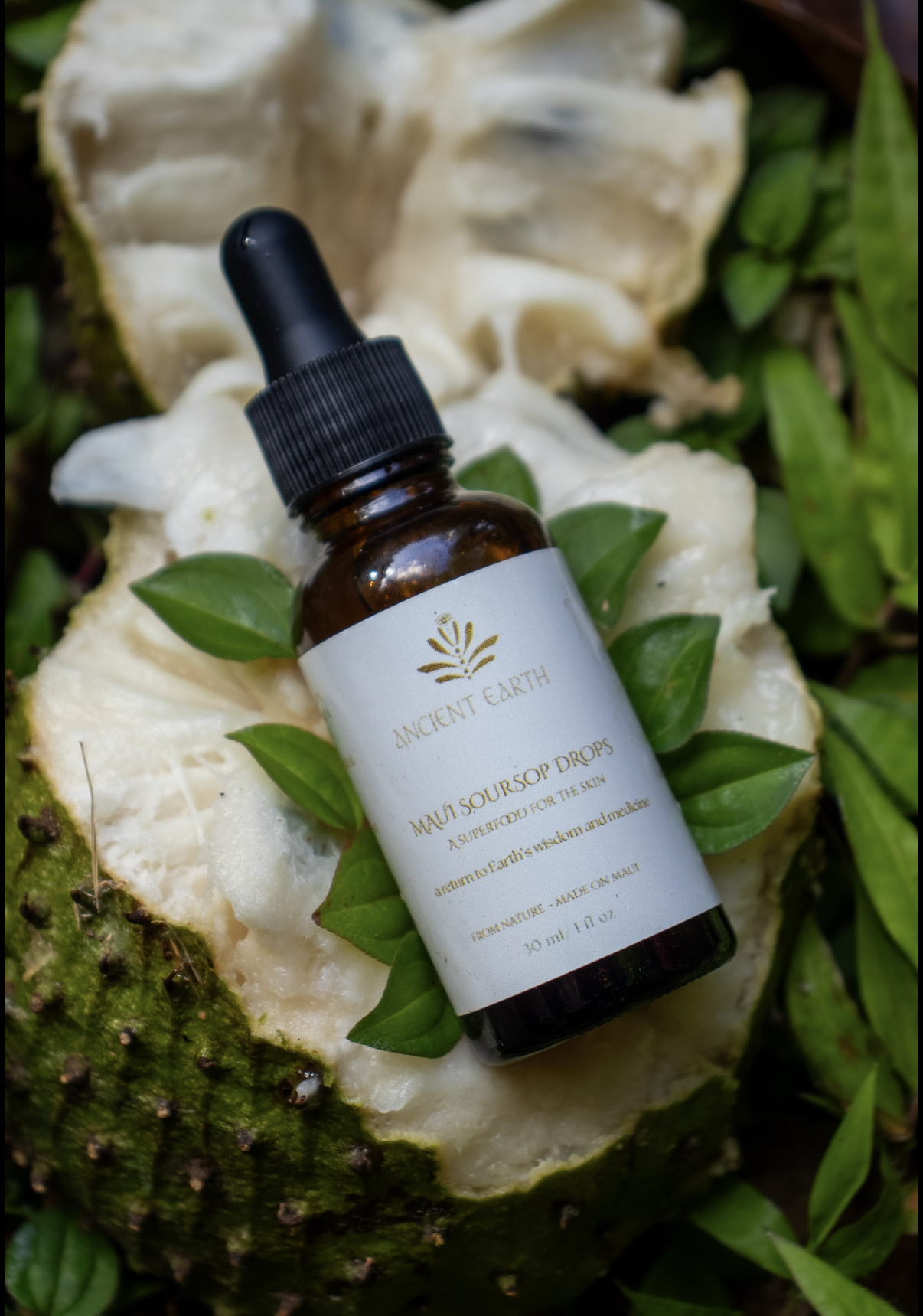 A small amber glass bottle of Ancient Earth Mauna Soursop Drops resting on a halved soursop fruit with green leaves in the background.