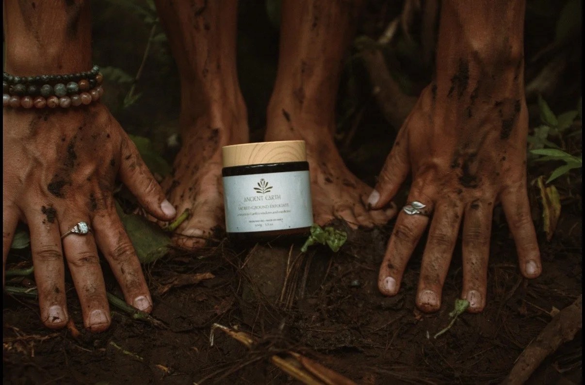 Two hands with dirt and rings on fingers touch the ground, surrounding a jar of Ancient Earth skincare product. The hands are adorned with necklaces and rings, with soil and greenery in the background.