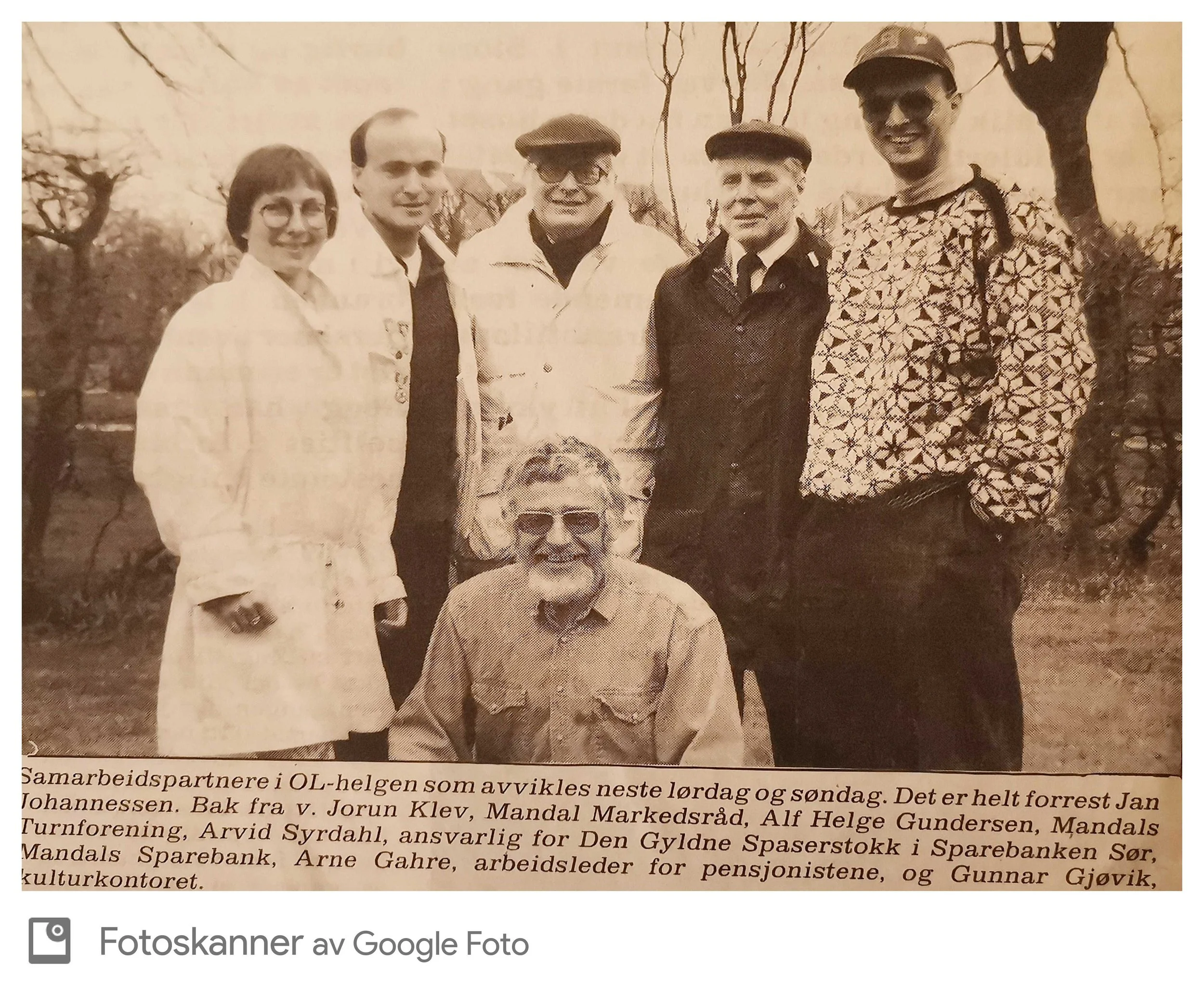 Seks personer som står utendørs foran trær, noen av dem smiler, og det er en hund foran dem. Fotografen er et eldre menneske med briller.