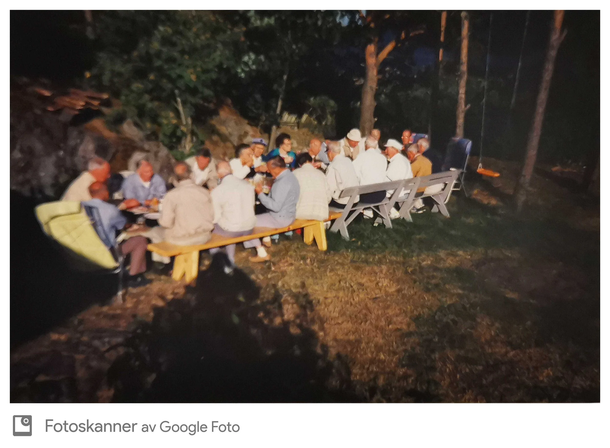 En gruppe mennesker sitter rundt en lang trebordsbord utendørs i skogen om kvelden, og ser ut til å nyte et samtale eller en aktivitet sammen.