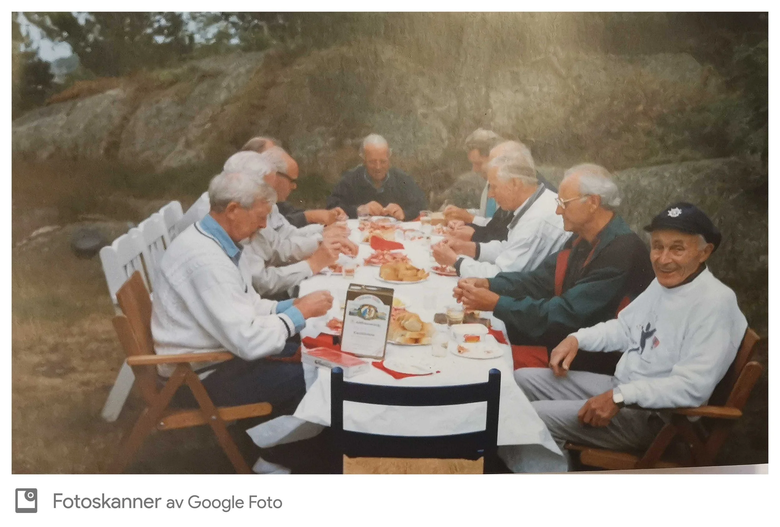 En gruppe eldre menn som sitter rundt et langbord utendørs og spiser middag. De ser ut til å ha en hyggelig sammenkomst.