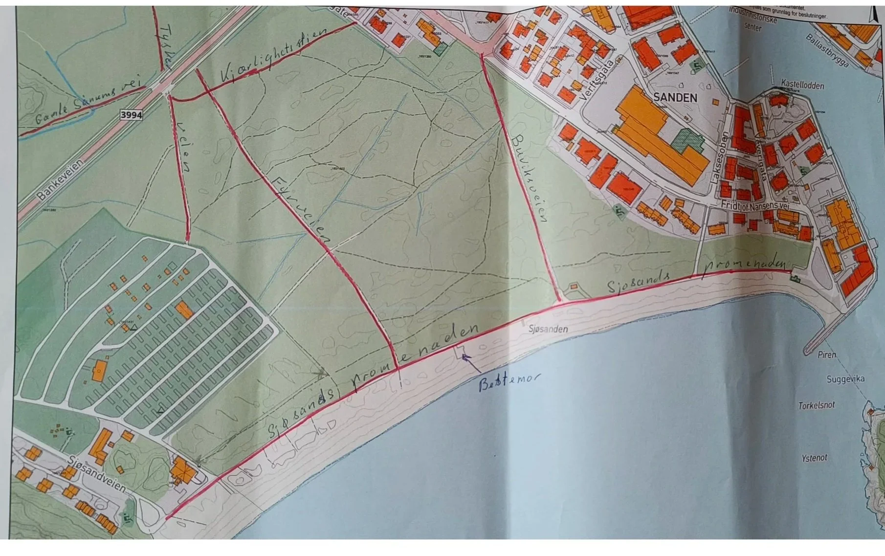 Kart over et område i Sjøsandskogen ved Sjøsanden Promonaden, med bygninger, veier og kystlinje langs sjøen, inkludert ulike farger og merkinger for terreng og strukturer.