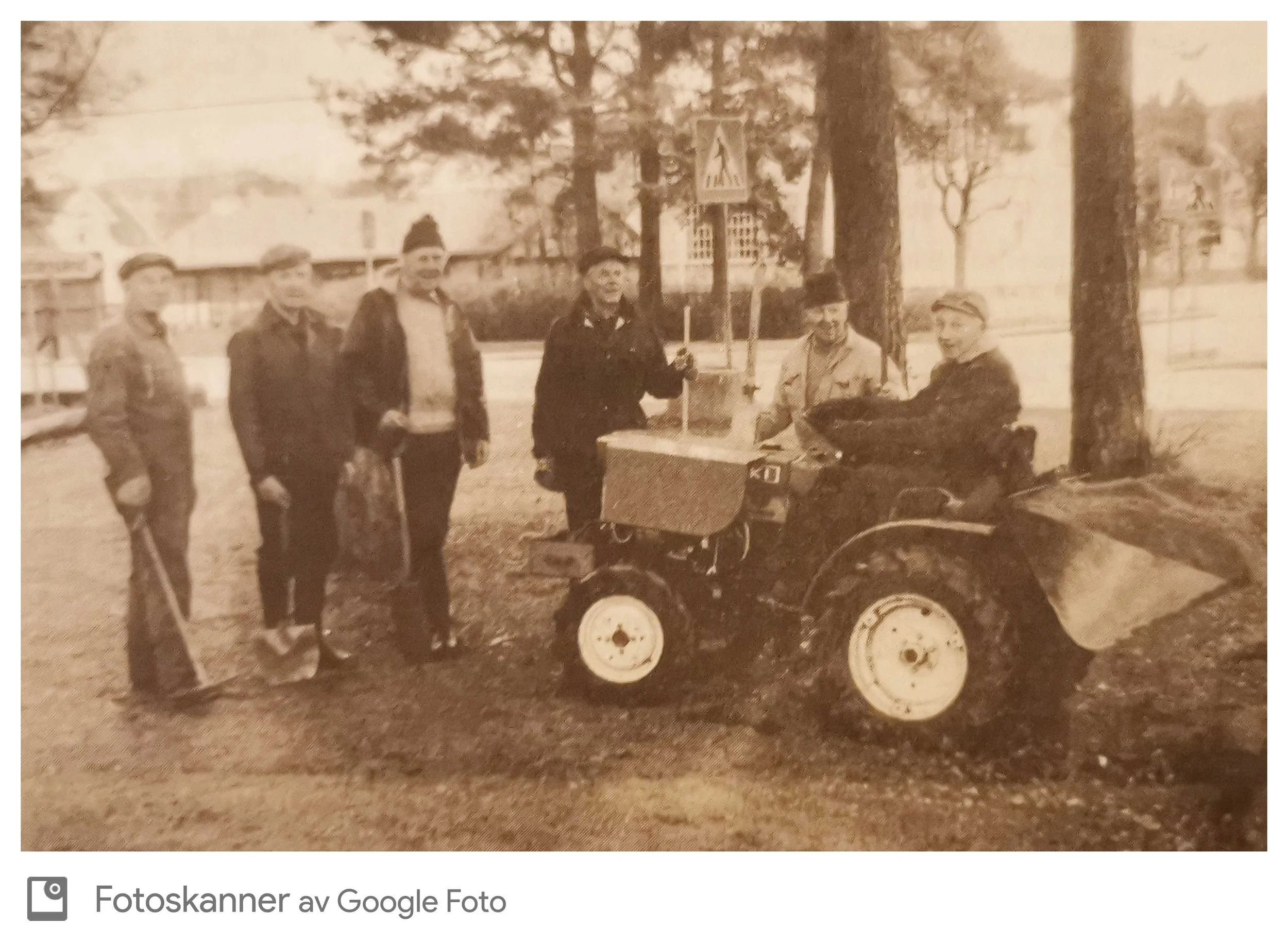 Seks personer står foran en traktor med fire hjul, som er parkert under noen trær. Alle ser ut til å være unge voksne og smilende, med noen iført vinterklær og noen med uniform. Bakgrunnen består av et gjerde og trær.