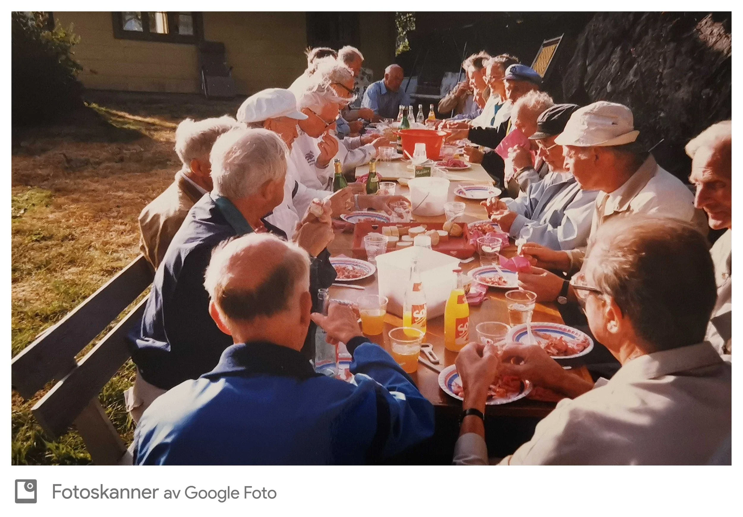 Dette er et bilde av en gruppe mennesker som spiser sammen utendørs på en lang bord. De ser ut til å nyte et festmåltid med pizza og drikke. Det er dag og det er solrike forhold.