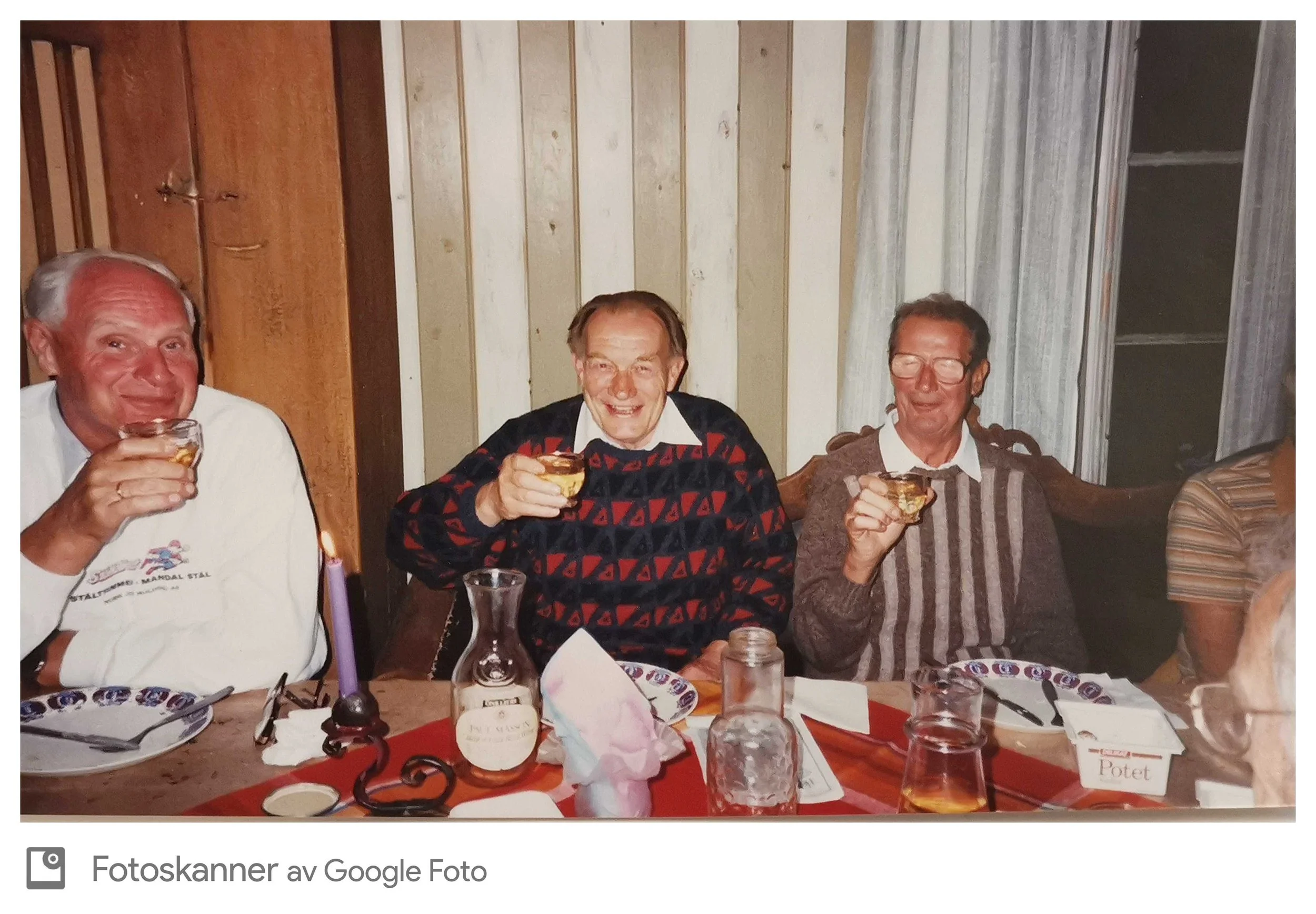 Tre menn som sitter ved et bord, smiler og holder hånden i luften, med drinker i hendene, foran et trehvitt panelvegger. Det er ulike mat- og drikkeobjekter på bordet, inkludert en bolle, en flaske, og en tallerken. To av mennene har briller, og alle