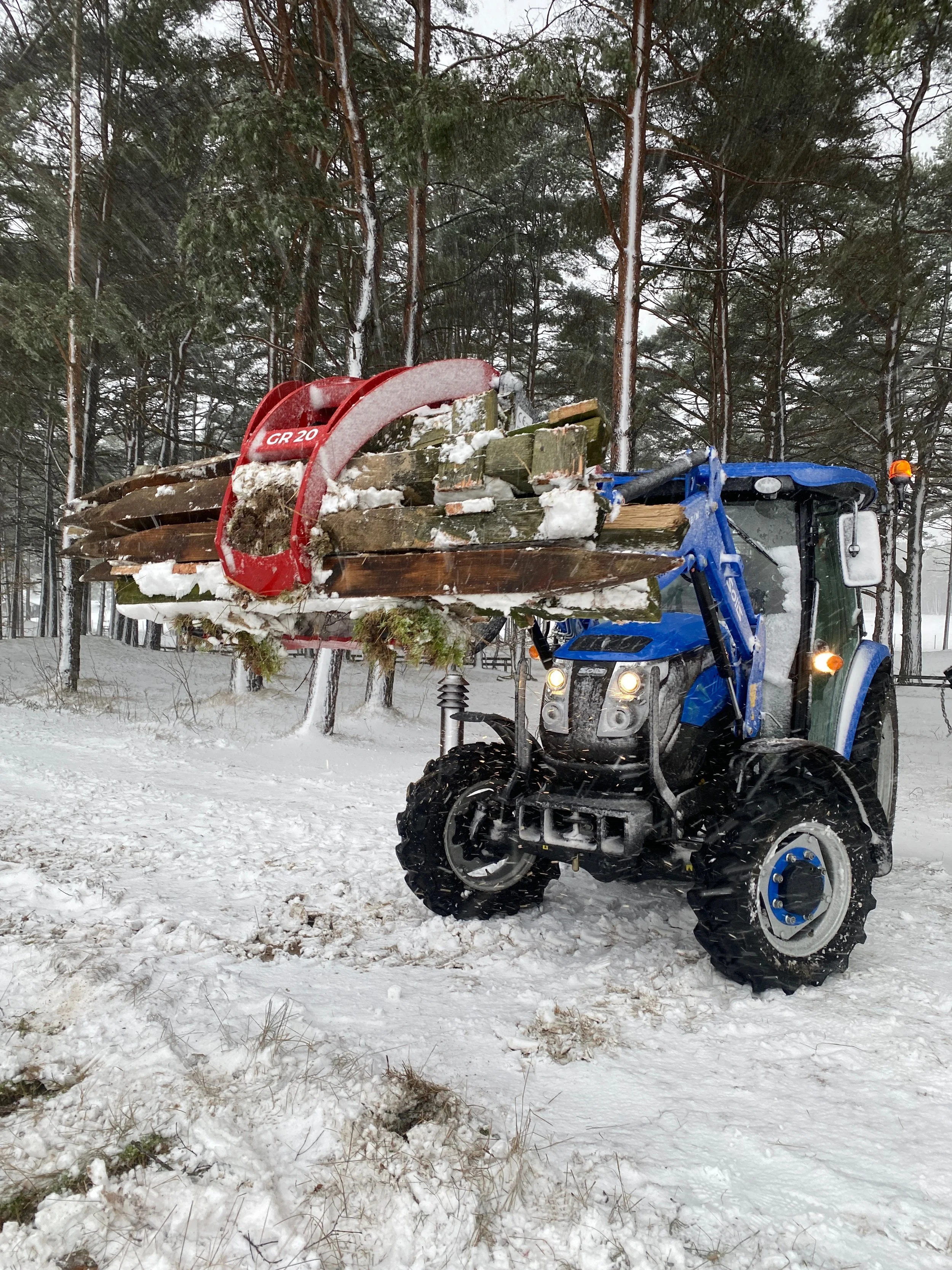 Bilde av en snødekket skog med en blå og svart traktor som har laster av trestammer på en redskap som kan brukes til å transportere dem.
