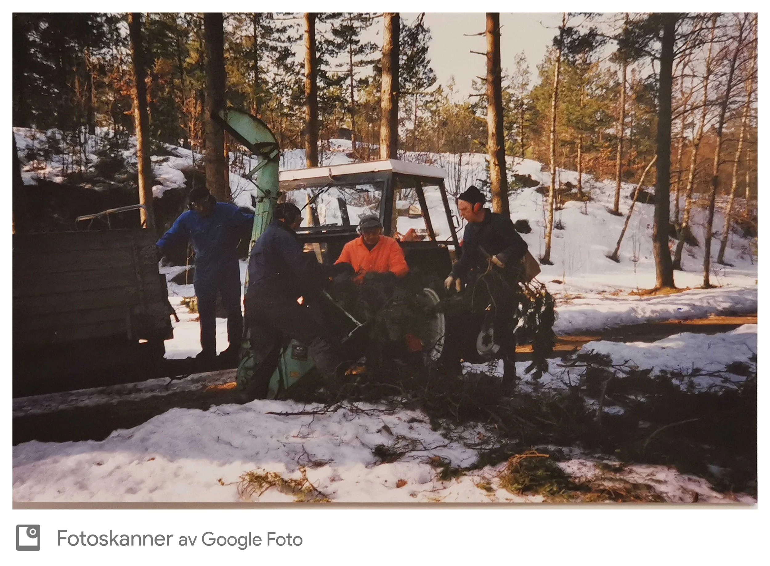 Personer i en skog om vinteren, med snø på bakken, som jobber med å rydde eller kutte ned trær. Flere personer er synlige, og det er en snømaskin eller traktor i bakgrunnen.