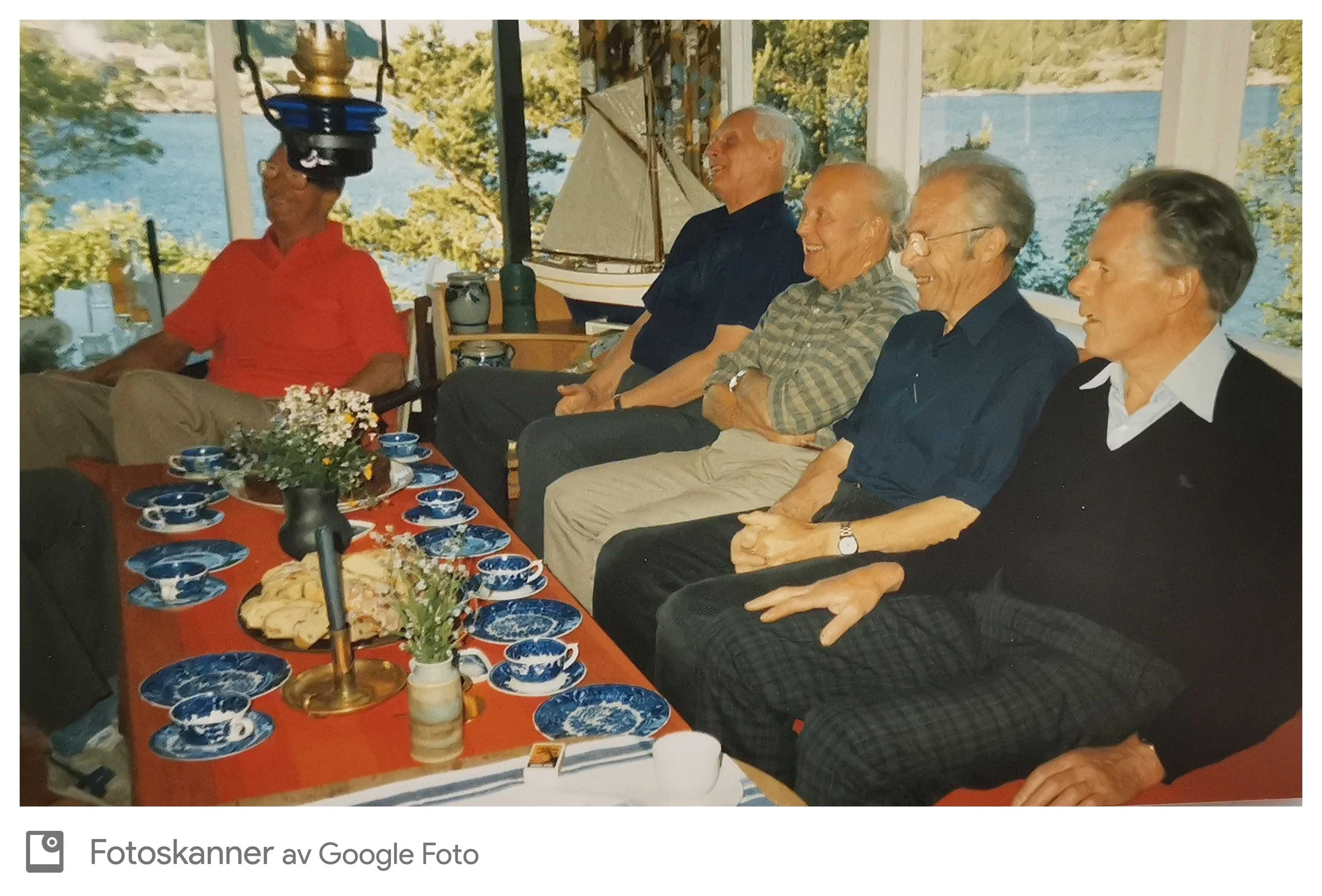 Fem eldre personer, tre menn og to kvinner, sitter side om side rundt et middagsbord dekket med blå og hvite serveringsfat, blomster og mat, inne i et lyst rom med store vinduer som viser en innsjø og trær utenfor. De ler og har det hyggelig.