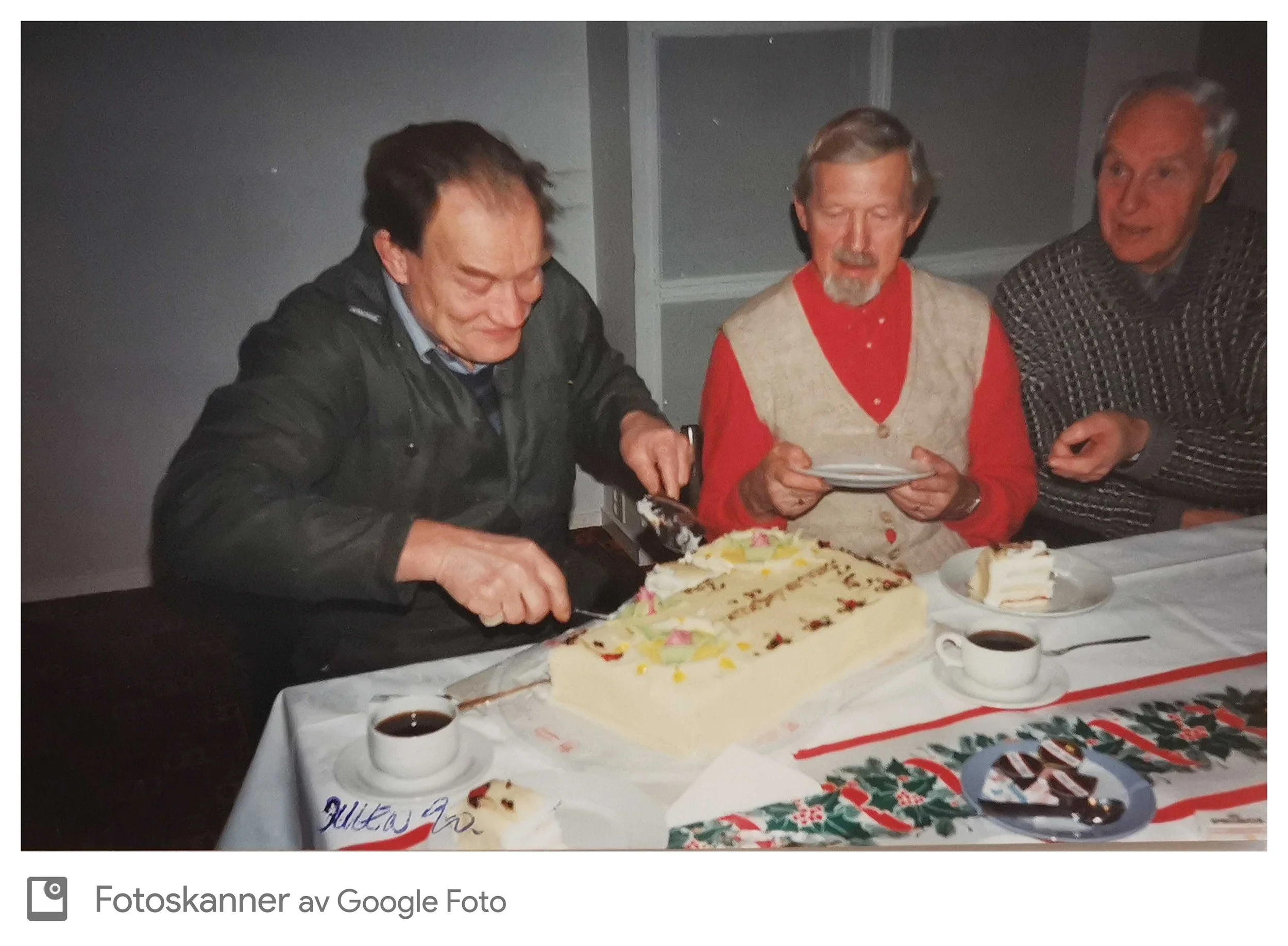 Tre menn har selskap rundt et bord med kake og drikke, og en av dem skjærer opp kaken, i en innendørs setting.