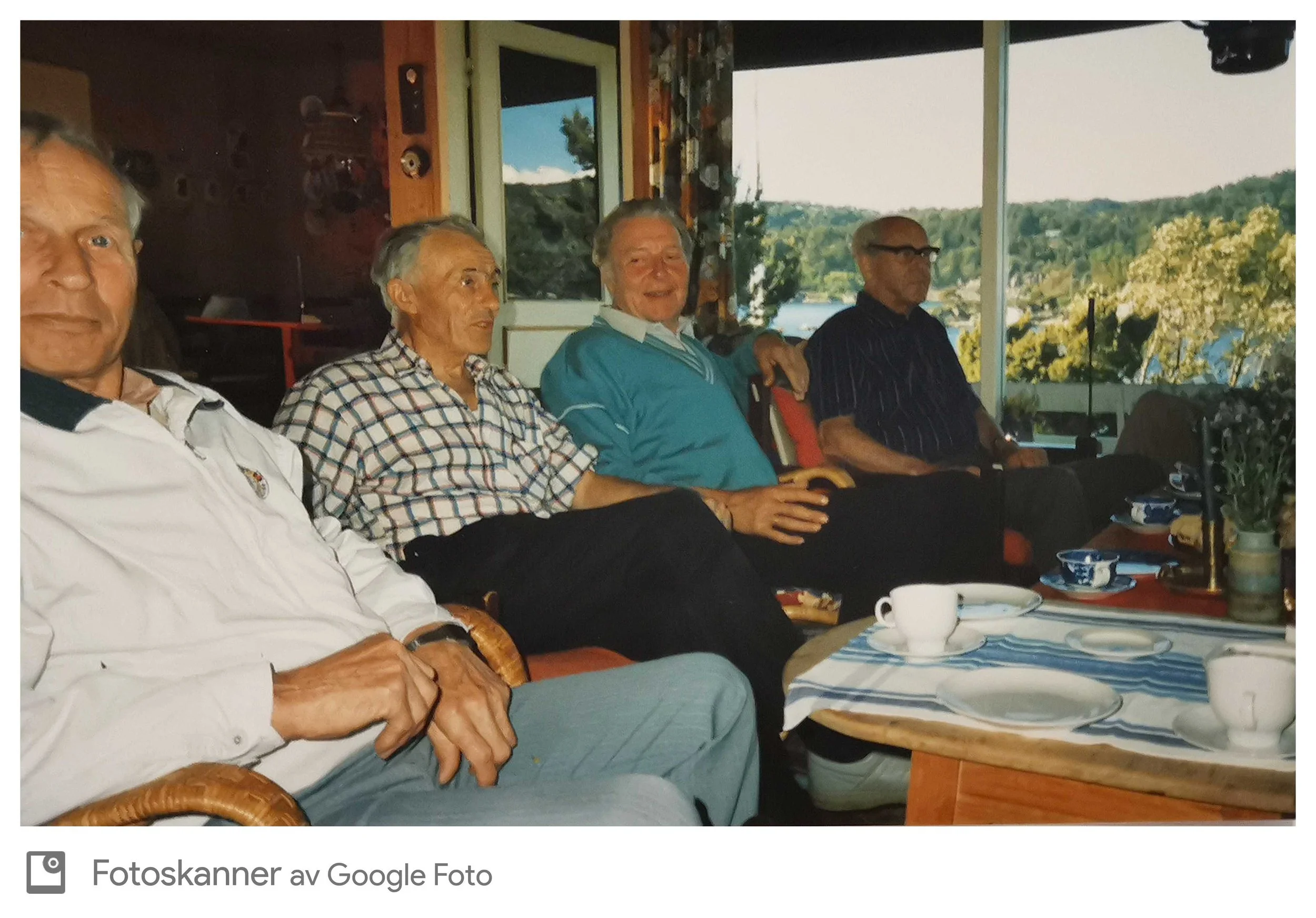 Fem eldre menn sitter på en veranda med utsikt over vann og grønne åser, og har på seg komfortable klær, noe som tyder på en samling eller sosiale sammenkomst.