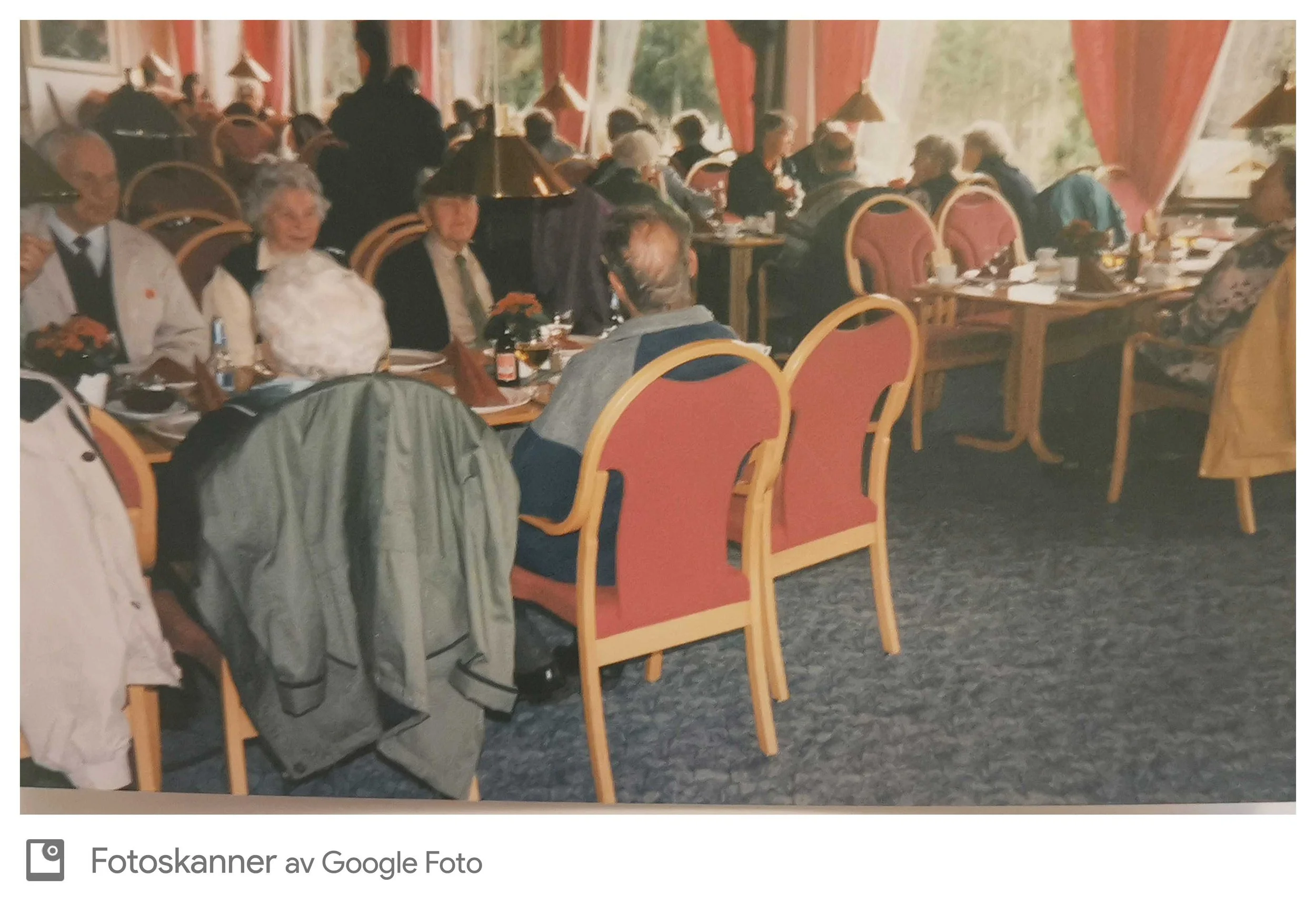 Innendørs restaurant med mange eldre mennesker som spiser og snakker. Borddekkene har tallerkener, kopper, og servise, og det er store vinduer med røde gardiner. Noen bruker briller, og det er lamper over bordene.