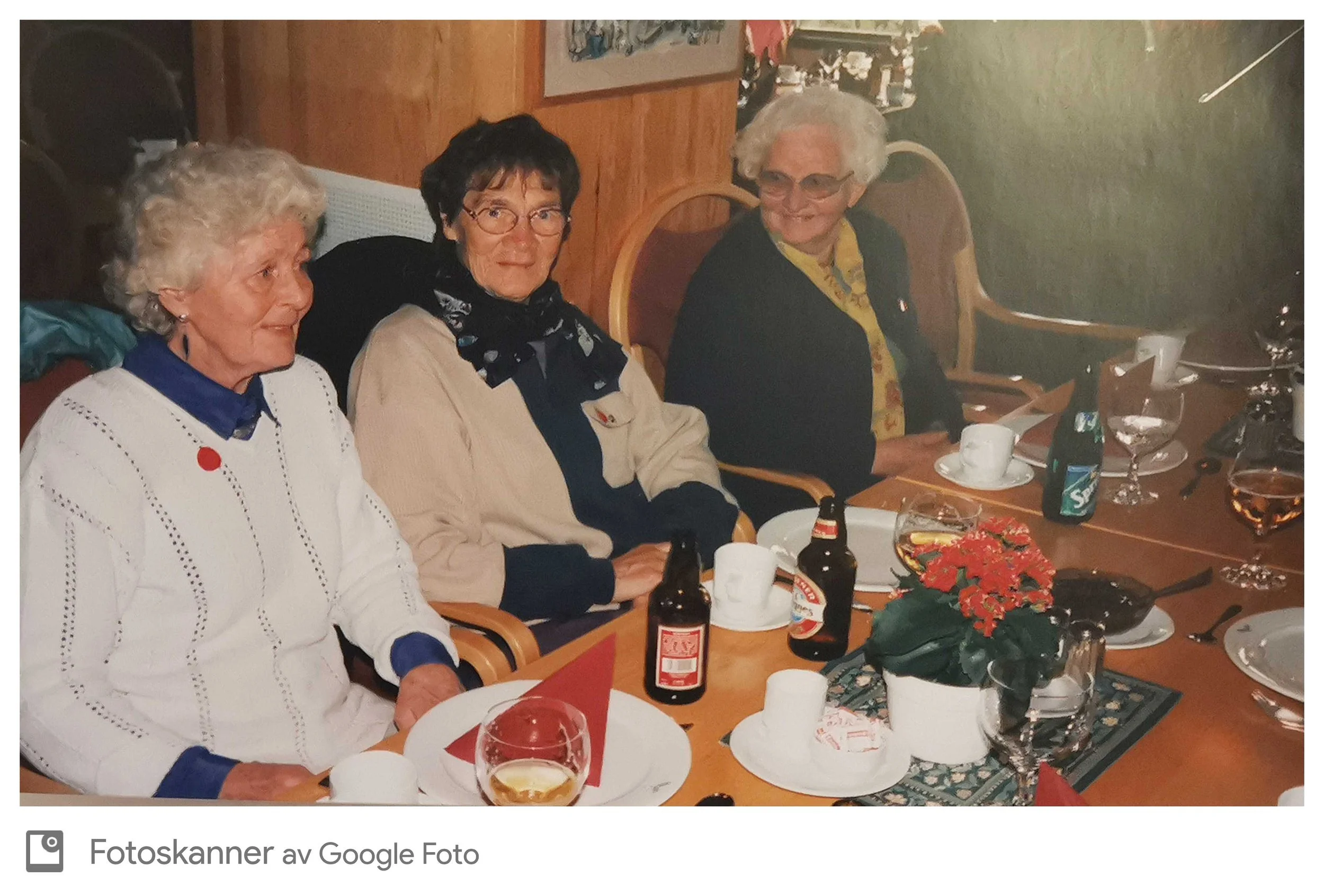 Tre eldre damer sitter rundt et spisebord med juledekorasjoner, drikkevarer og mat. De ser ut til å ha en hyggelig samling.