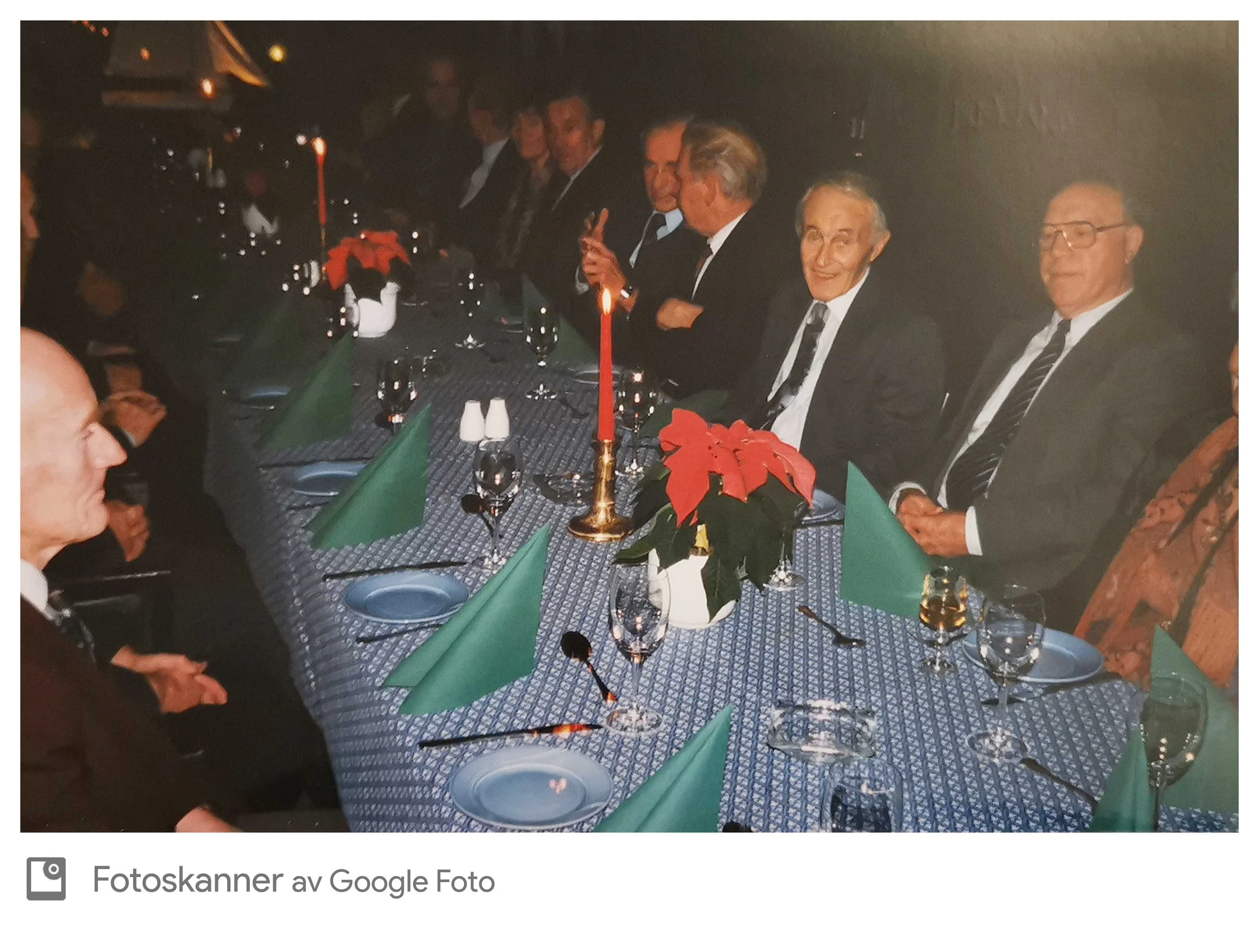 Gruppemennesker som sitter rundt et langbord dekket med en bordduk, med julepynt og lys, i en festlig setting.
