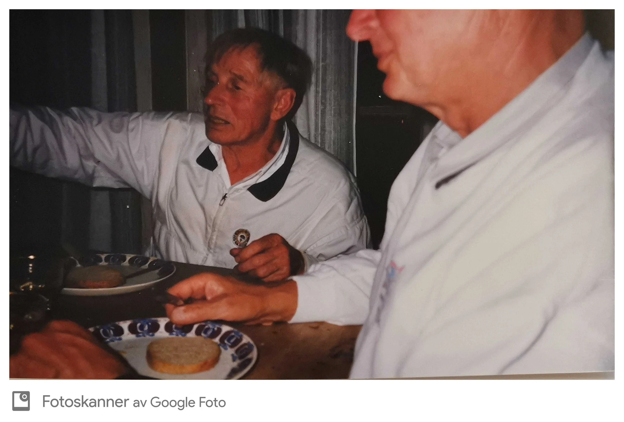 To personer, begge i hvite klær, som spiser hamburgere sammen. En person er tydelig synlig med mørkt hår og sier noe, mens den andre er mer avbildet i profil uten synlig ansikt.