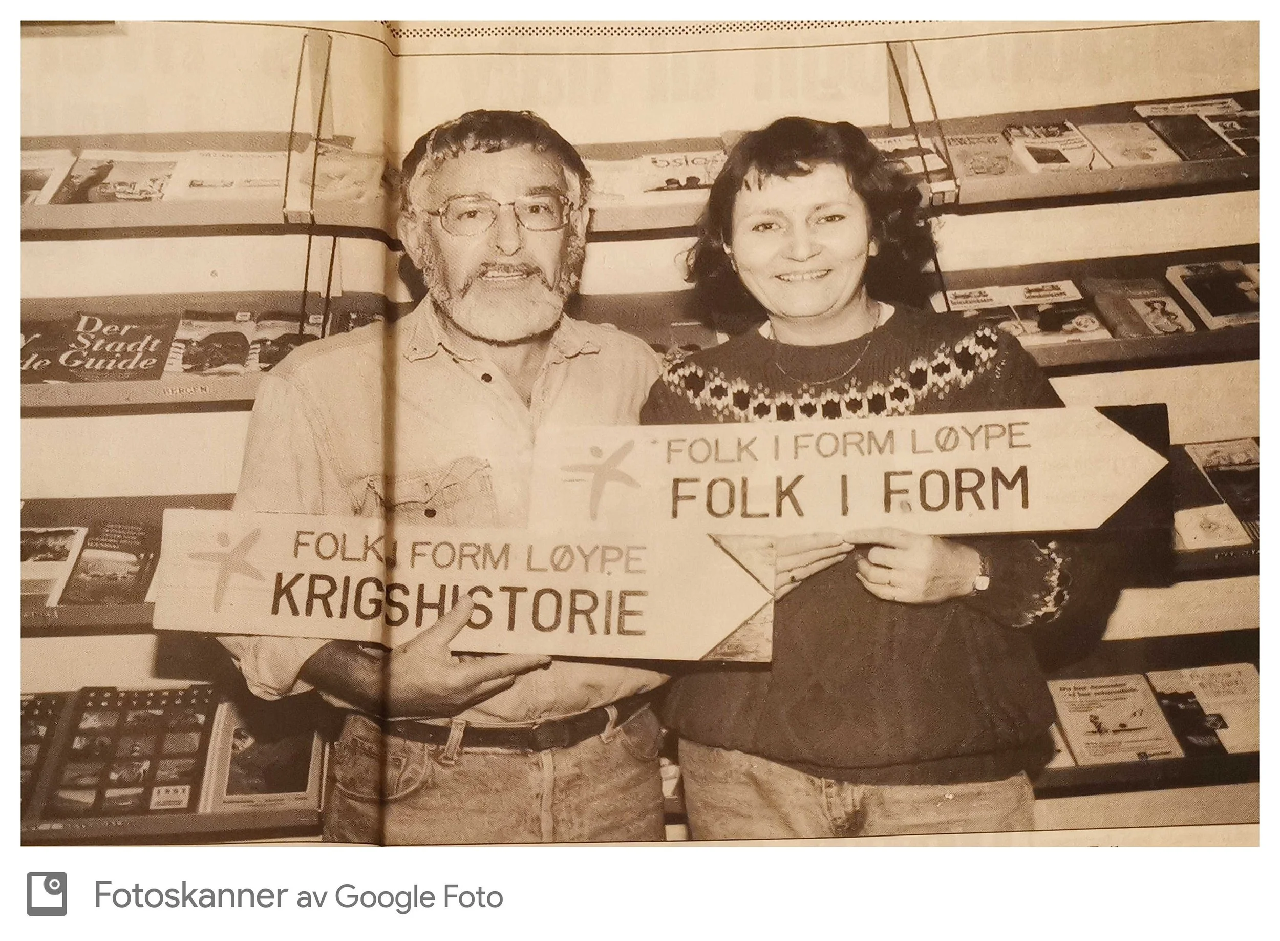En svart-hvitt foto av en mann og en kvinne som smiler og holder en plakat med teksten 'FOLK I FORM LØYPE' og andre ord som 'KRIGSHISTORIE'. De står foran hyller med bøker og magasiner.